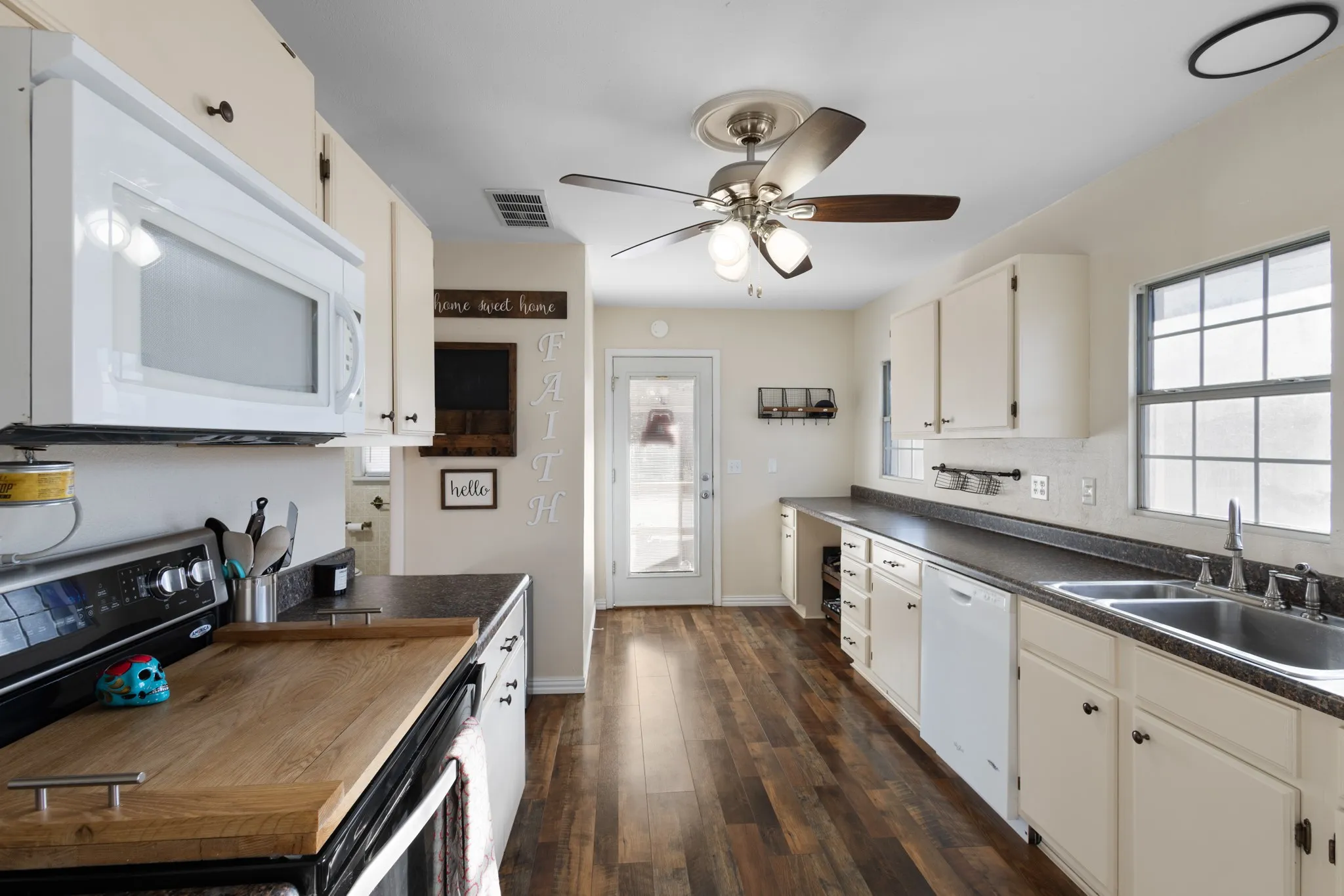 White cabinetry with dark hardware, dark countertops, wood-finish flooring, stainless steel sink, and white appliances