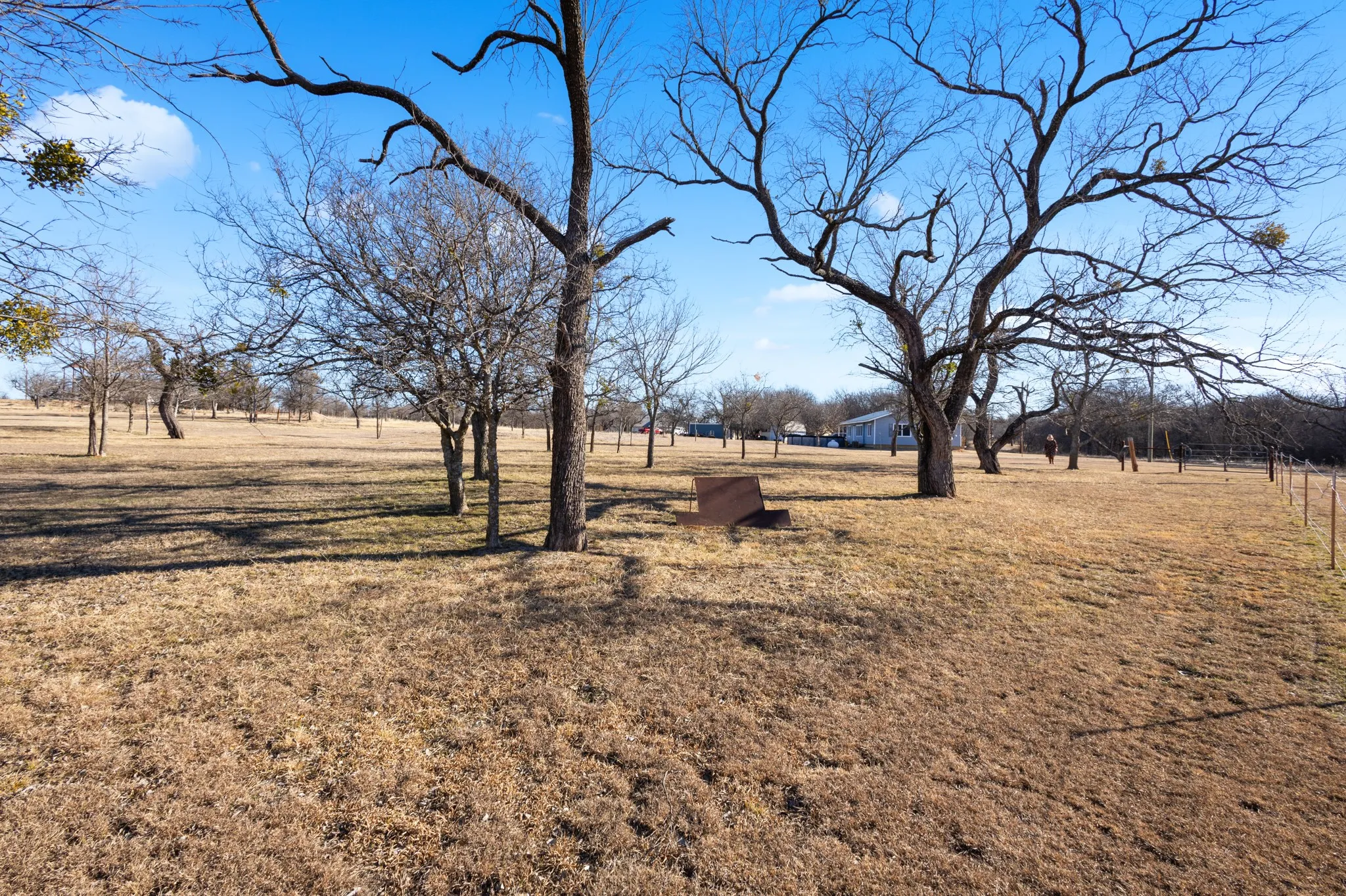 Expansive property with mature trees and open land, featuring a distant single-story structure and a partial fence line