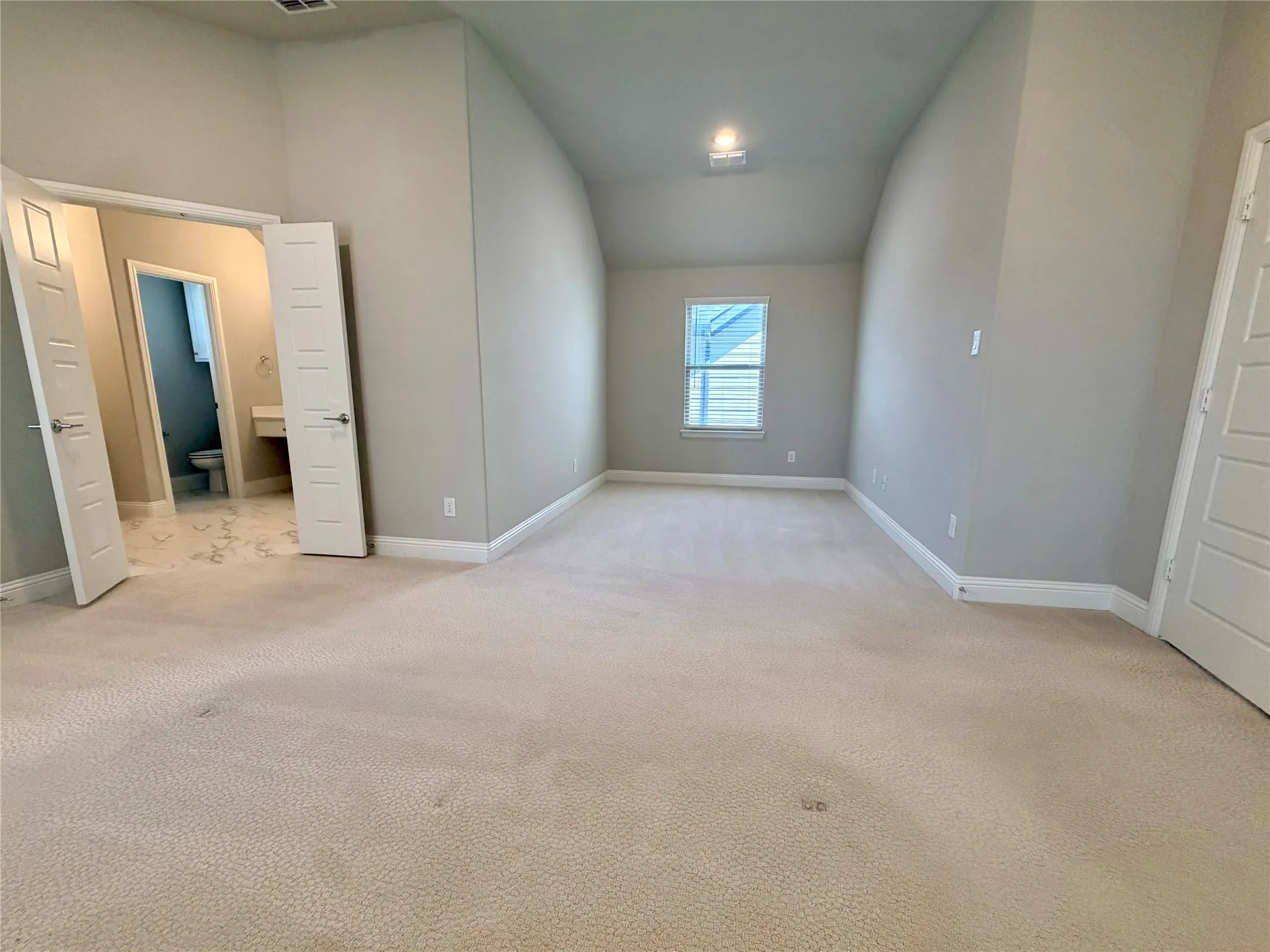 Spacious room with neutral carpeting, light gray wall paint, a single window, and an adjacent bathroom featuring a vanity