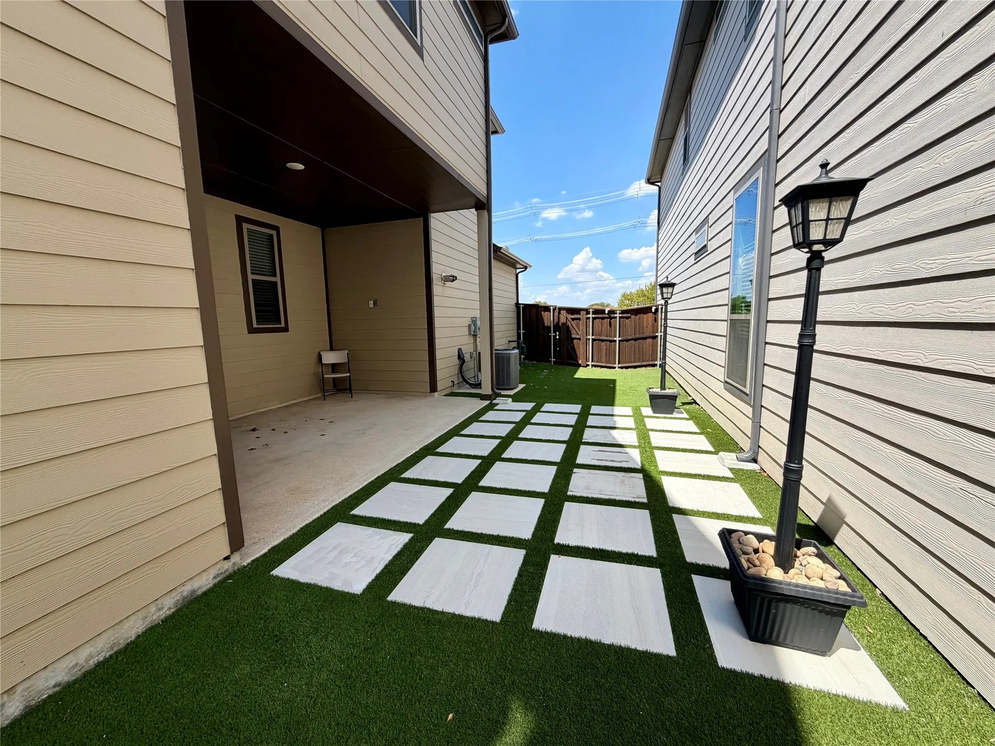 Covered concrete patio with recessed lighting, artificial turf, square paver walkway, and exterior wall lanterns