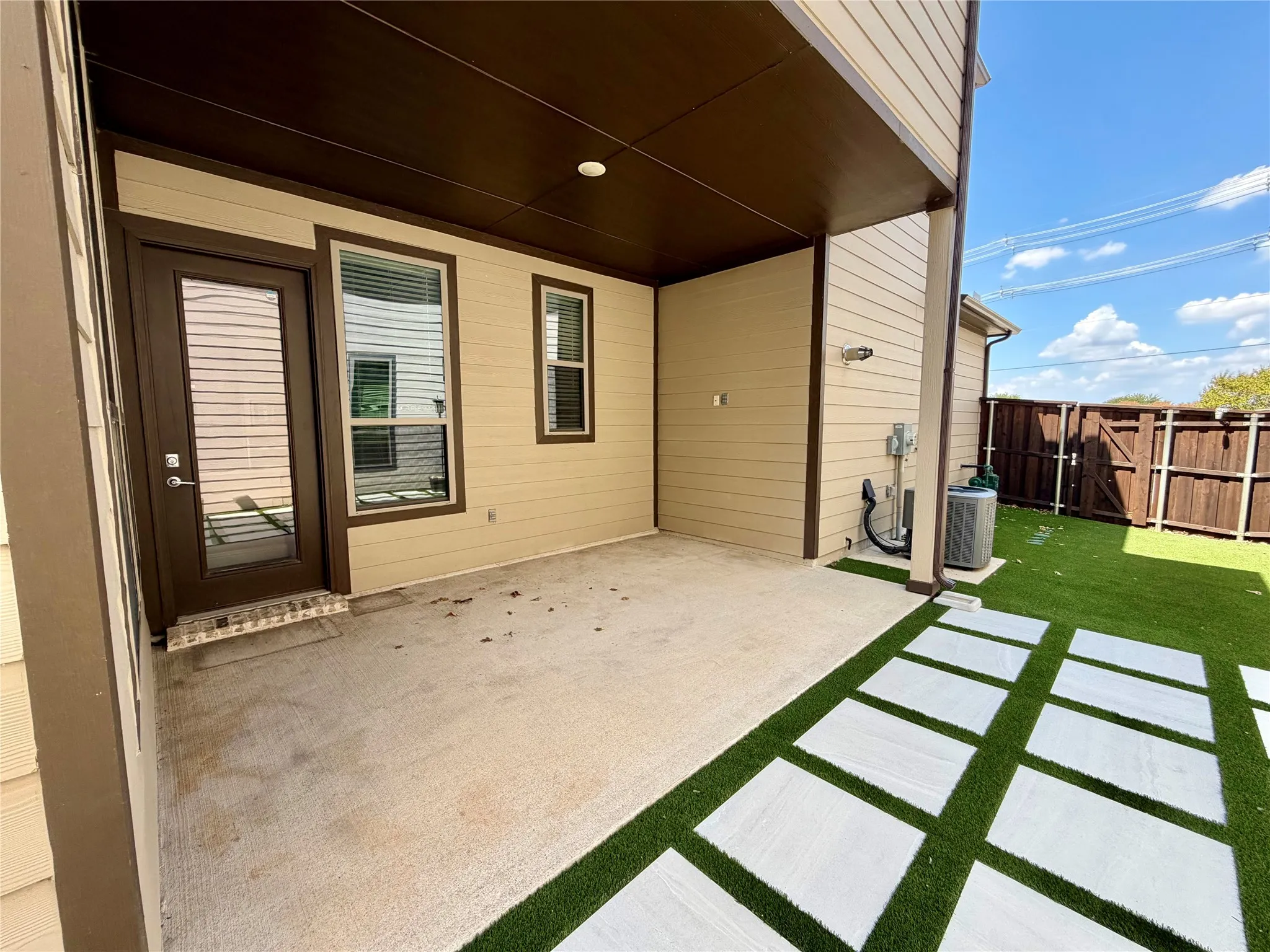 Covered concrete patio with a full-light door, two windows with blinds, and an artificial turf yard with paver stepping stones