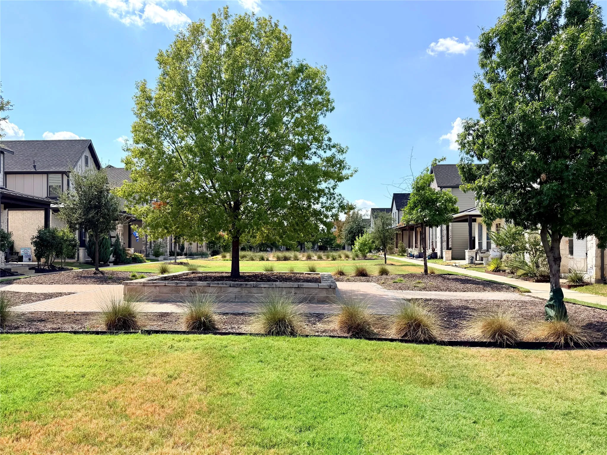 Community green space featuring mature trees, landscaped beds, and a paver pathway system