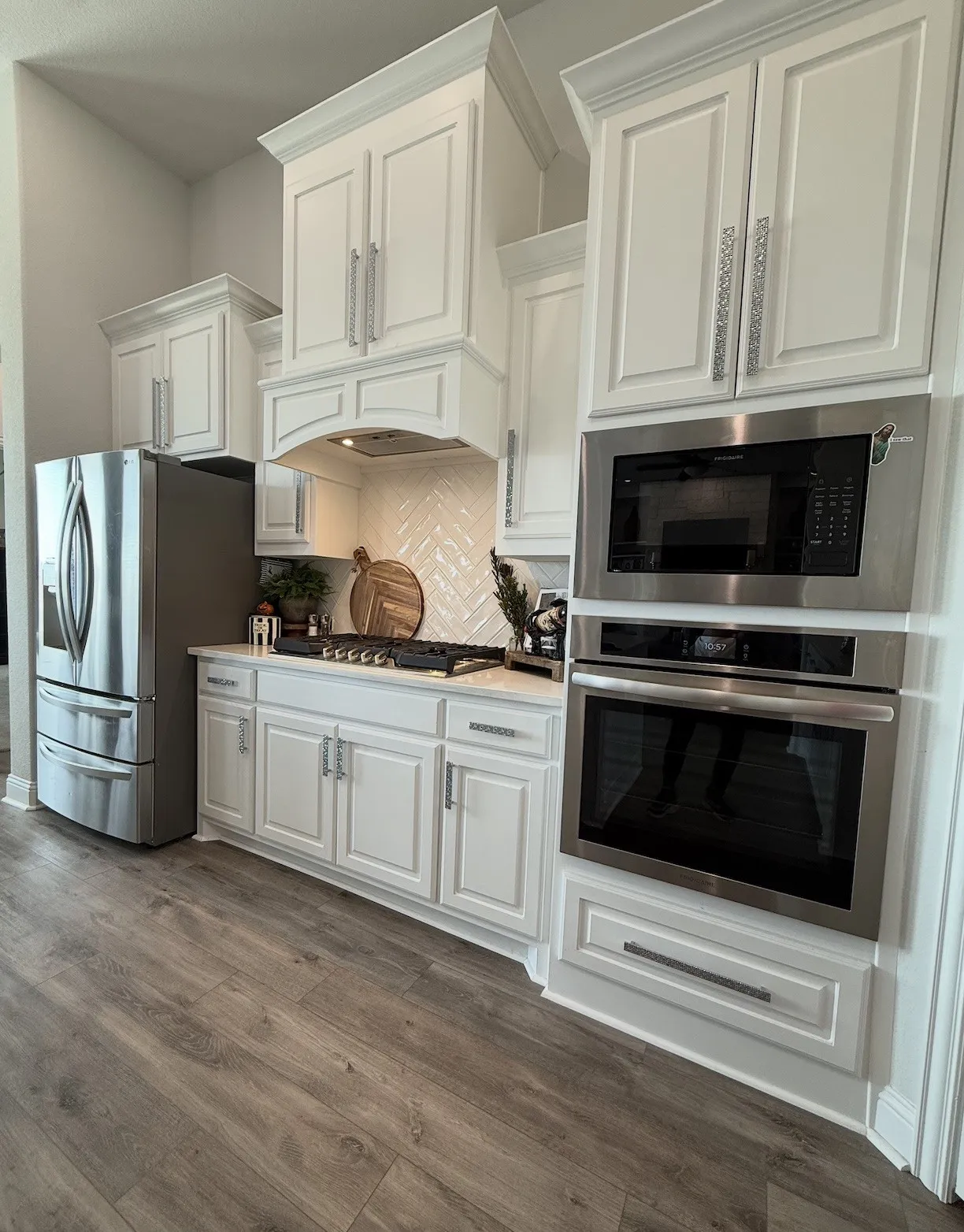 White cabinetry with crown molding, stainless steel appliances, herringbone tile backsplash, integrated range hood, and wood-finish flooring