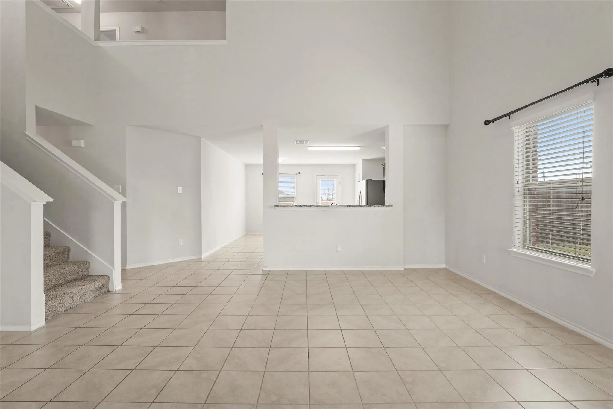 Open concept living area with high ceilings, featuring tile flooring, a staircase with carpeted treads, and an integrated kitchen pass-through