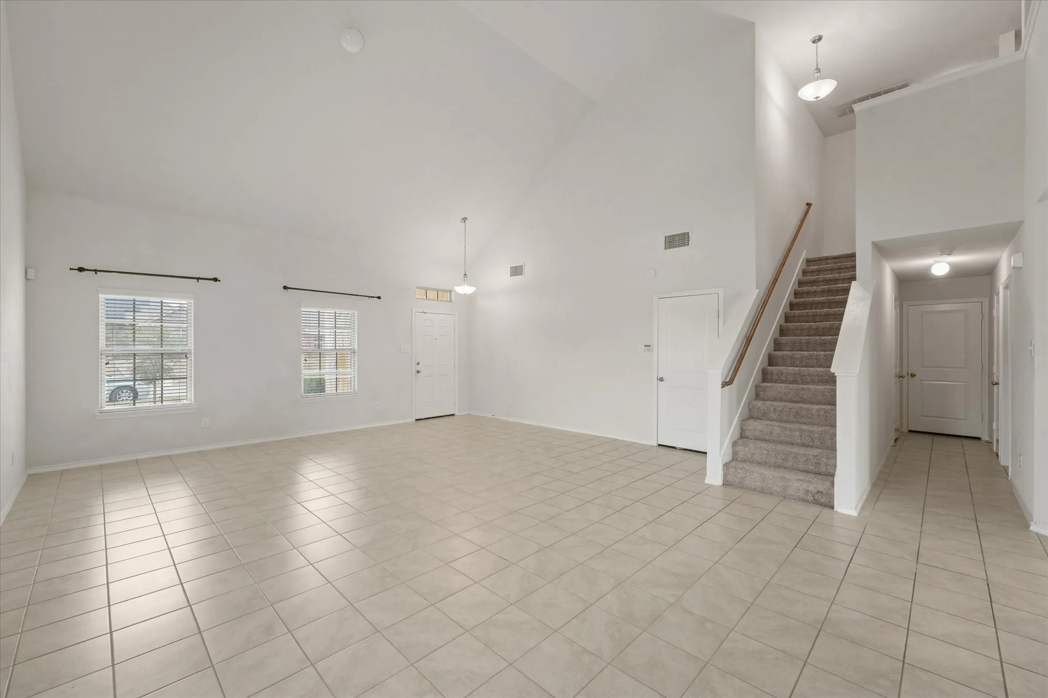 Vaulted ceilings, neutral tile flooring, two windows with horizontal blinds, and a carpeted staircase with a wood handrail