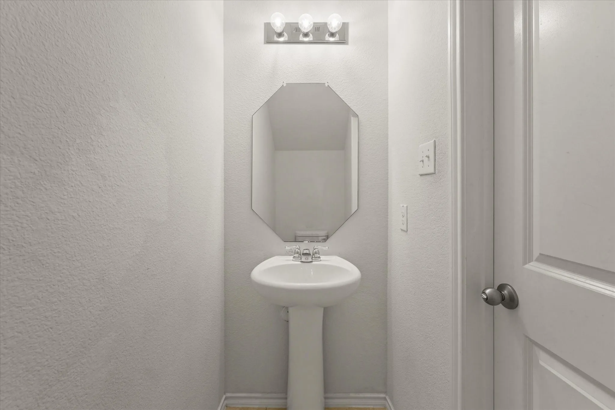 Pedestal sink with chrome faucet, octagonal mirror, and a three-light vanity fixture