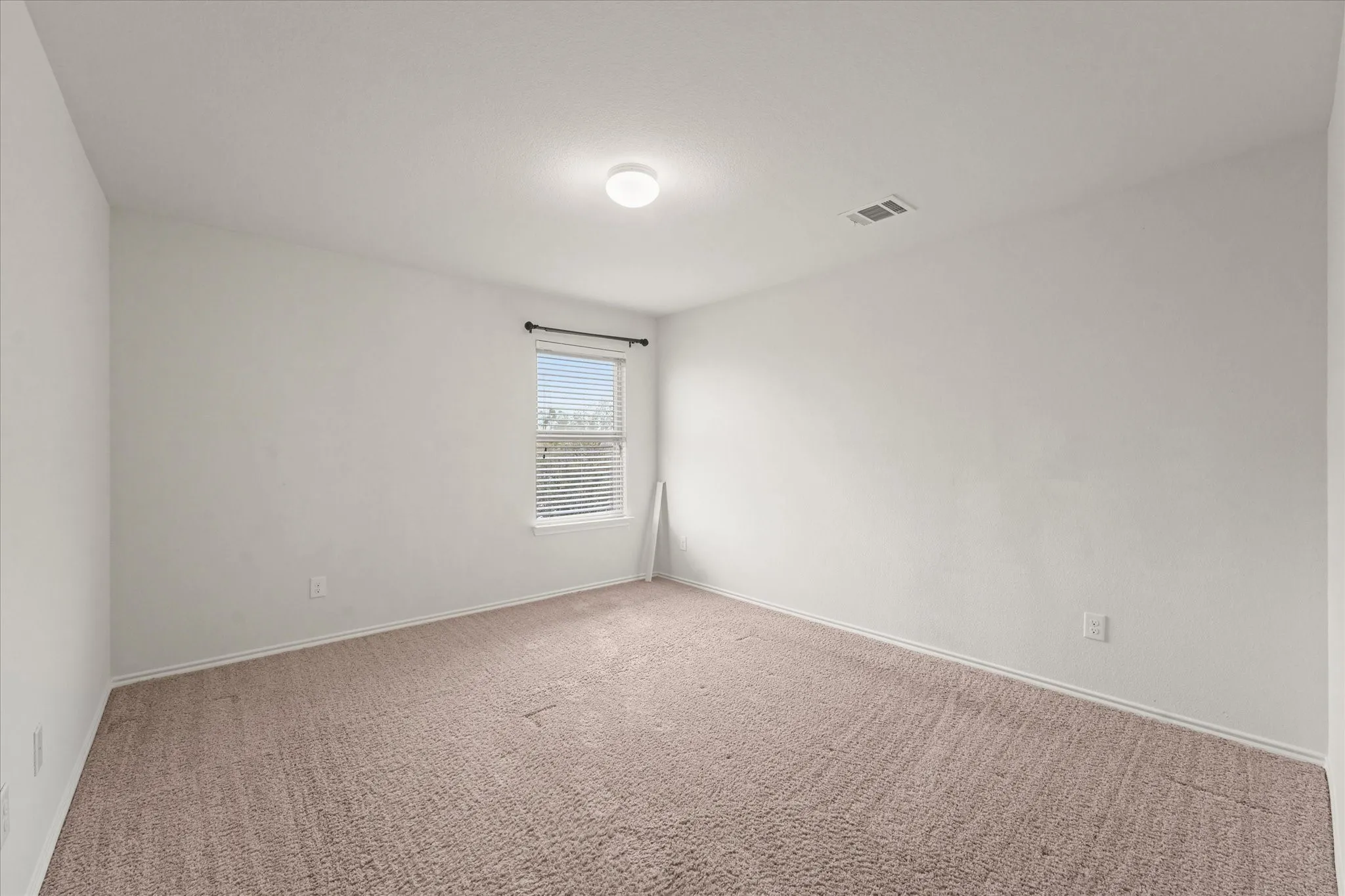 Carpeted flooring, single window with blinds, ceiling-mounted light fixture, white walls, and a wall outlet