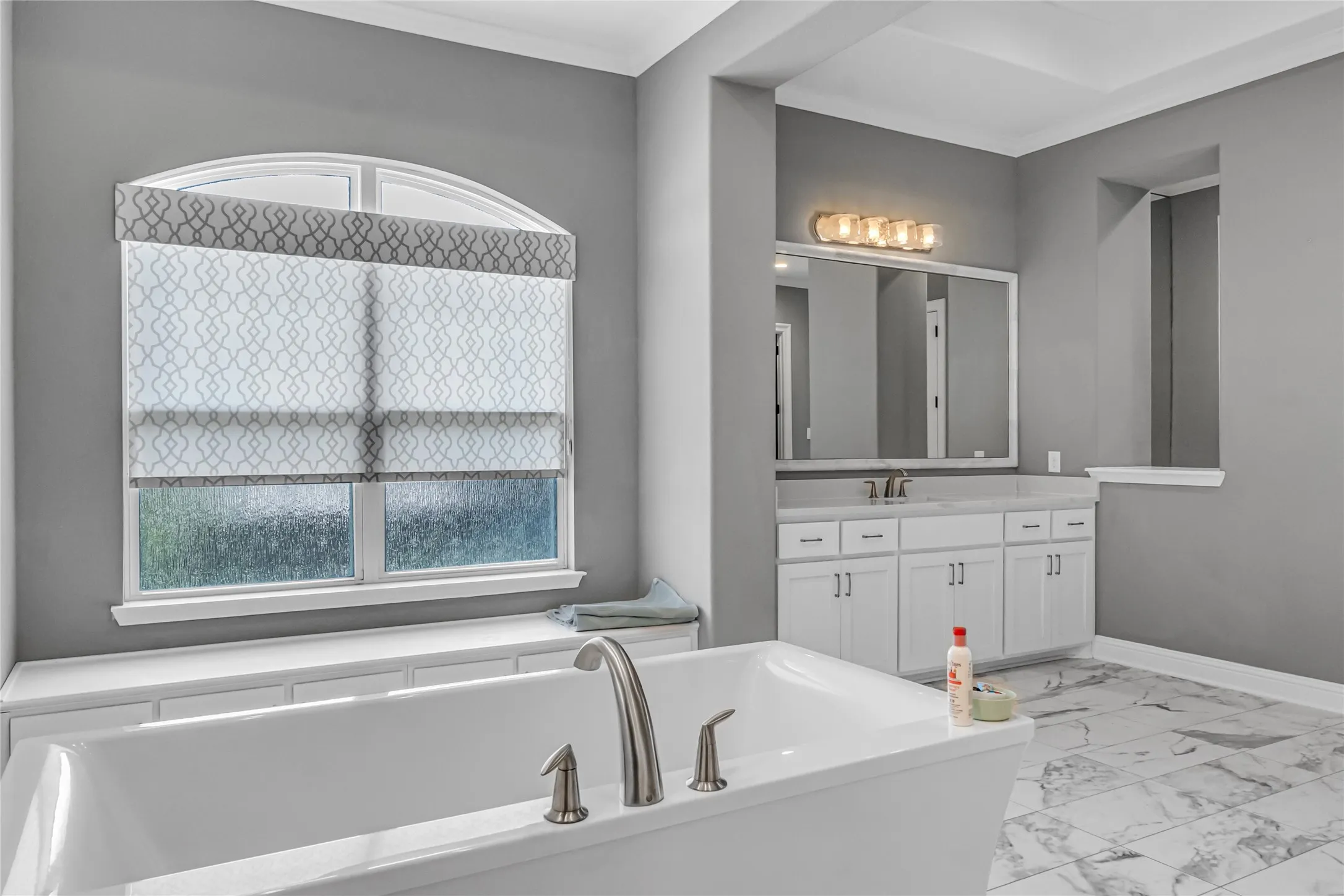 Freestanding soaking tub with brushed nickel fixtures, arched window, double vanity with white cabinetry, framed mirror, and tile flooring