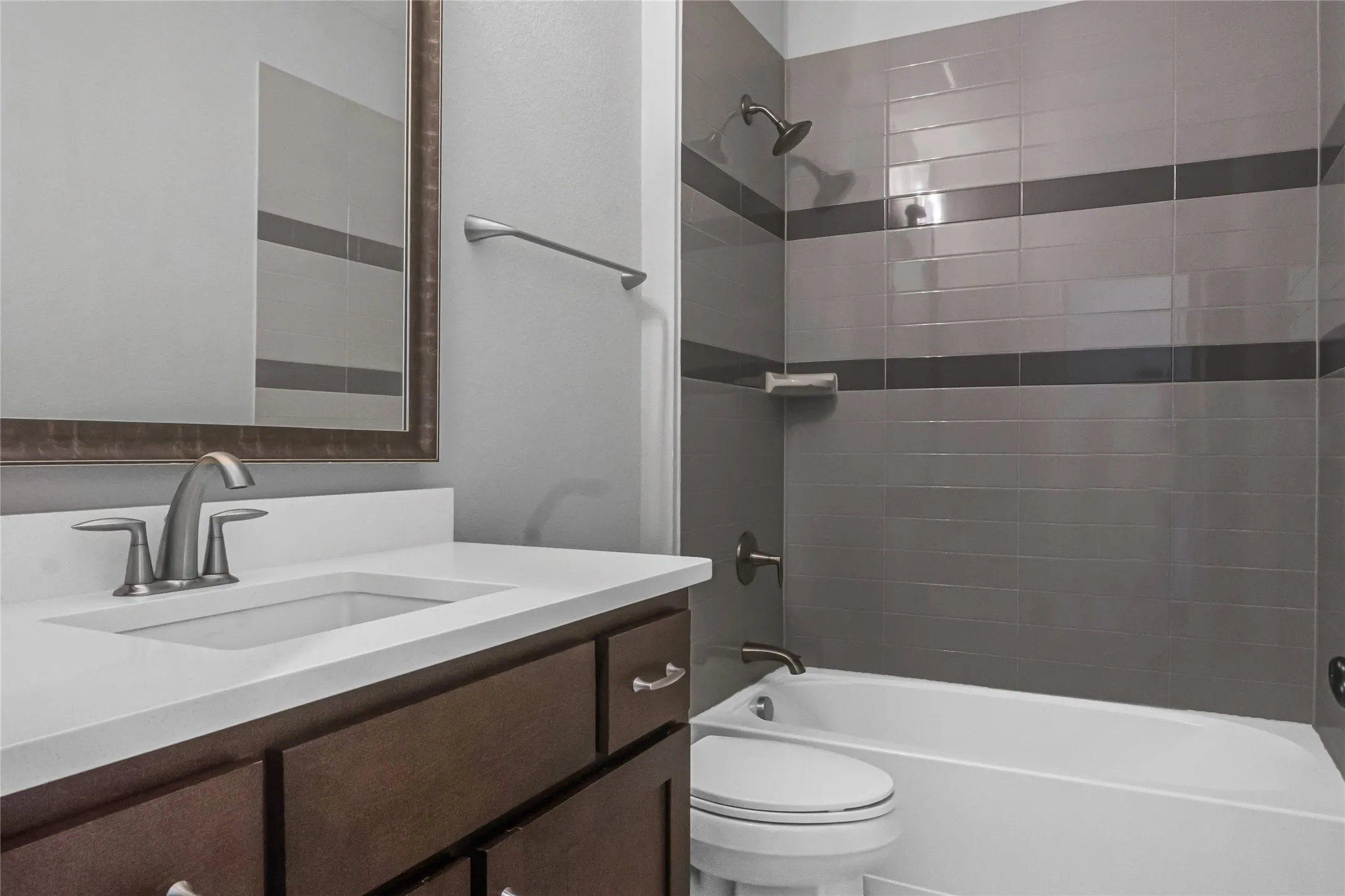 White countertop with integrated sink, dark wood-finish vanity, brushed nickel fixtures, subway tile shower surround with contrasting accent bands, and a white bathtub