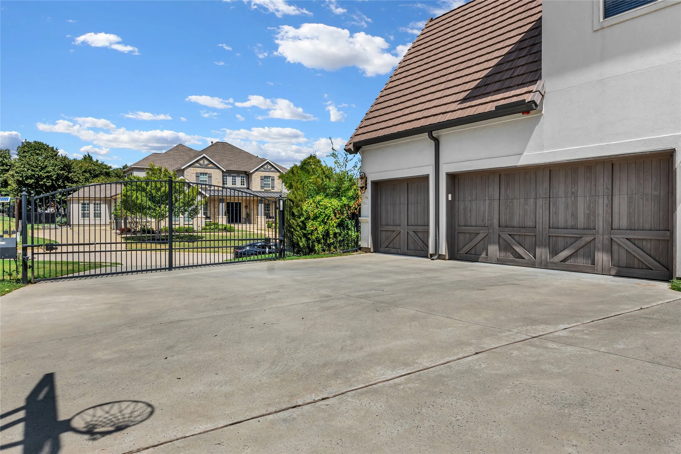 Concrete driveway with a basketball hoop shadow, wrought iron gate, stucco exterior, tile roof, and wooden garage doors