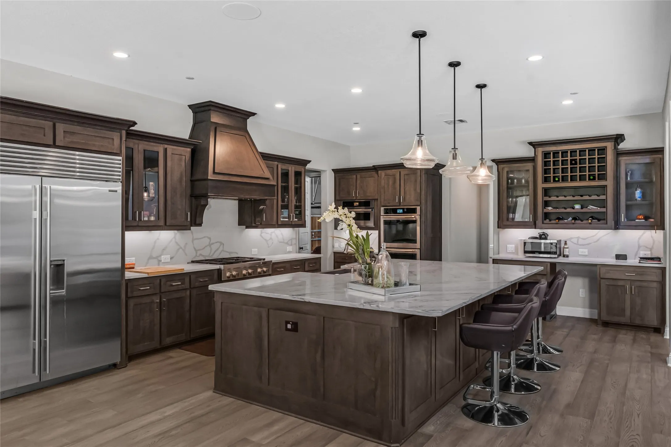 Gourmet kitchen featuring a large central island with seating, stainless steel appliances, custom wood cabinetry, a built-in wine rack, and wood-finish flooring