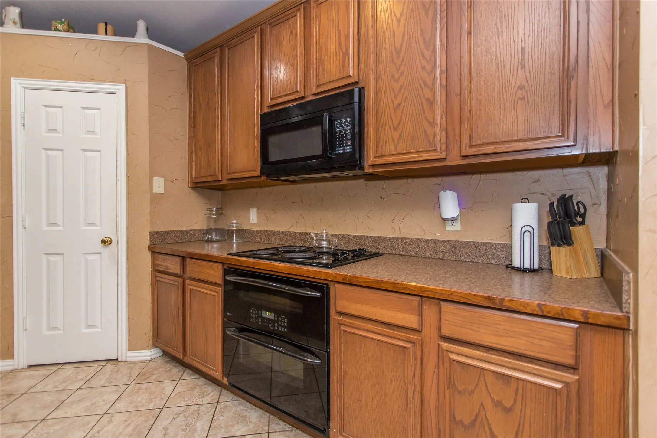 Wood-finish cabinetry, built-in black appliances, textured backsplash, and stone-finish countertops