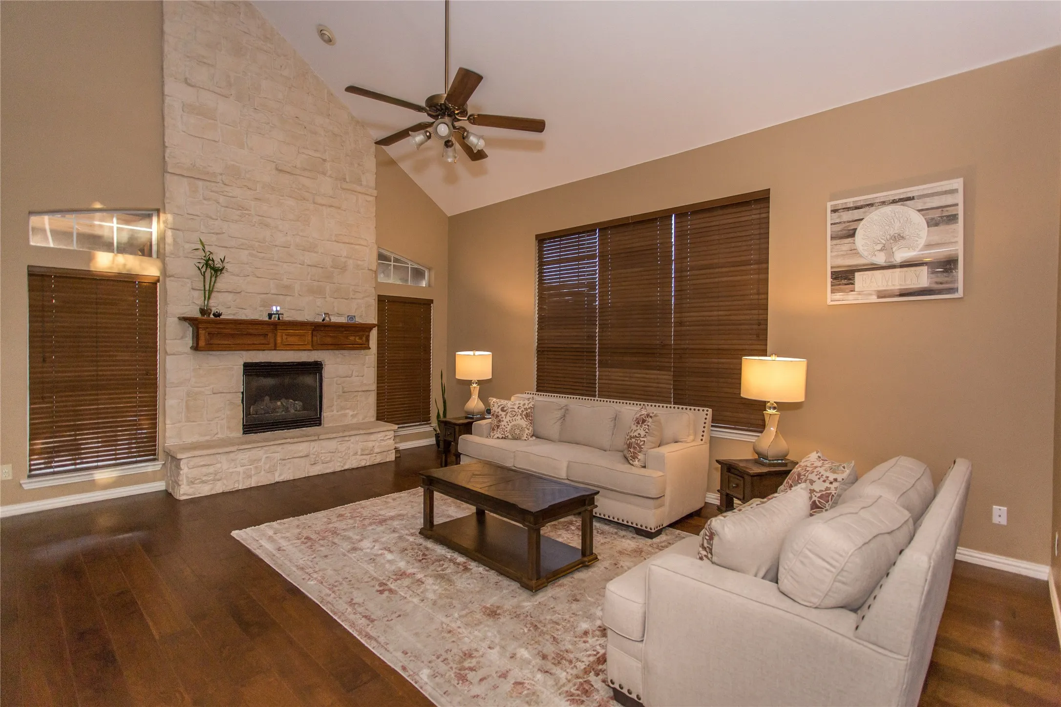 Vaulted ceiling with a ceiling fan, stone fireplace with a wood mantle, hardwood flooring, and multiple windows with wood blinds