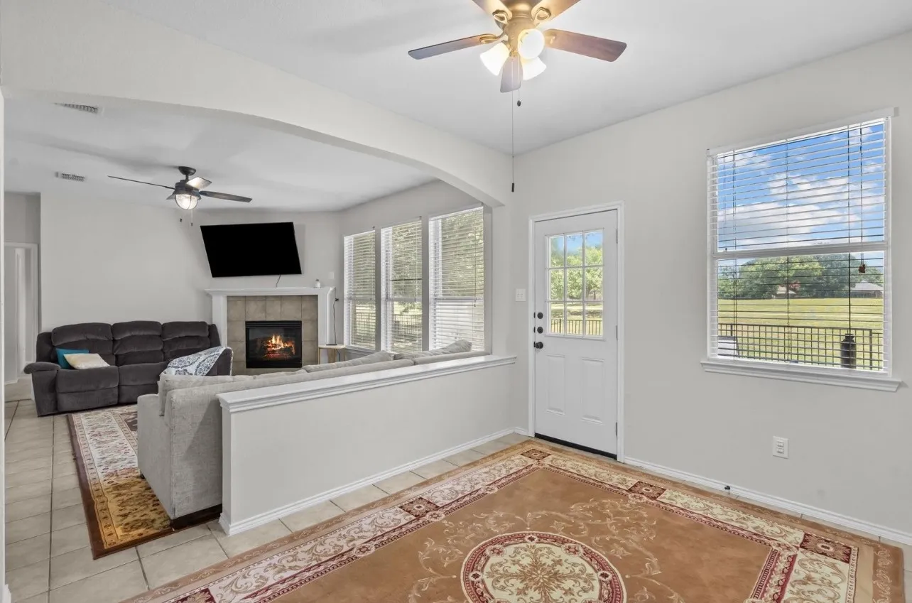 Bright sunroom with gleaming wood-finish flooring, decorative area rug, ceiling fan with integrated lighting, and expansive windows framing pastoral landscape views