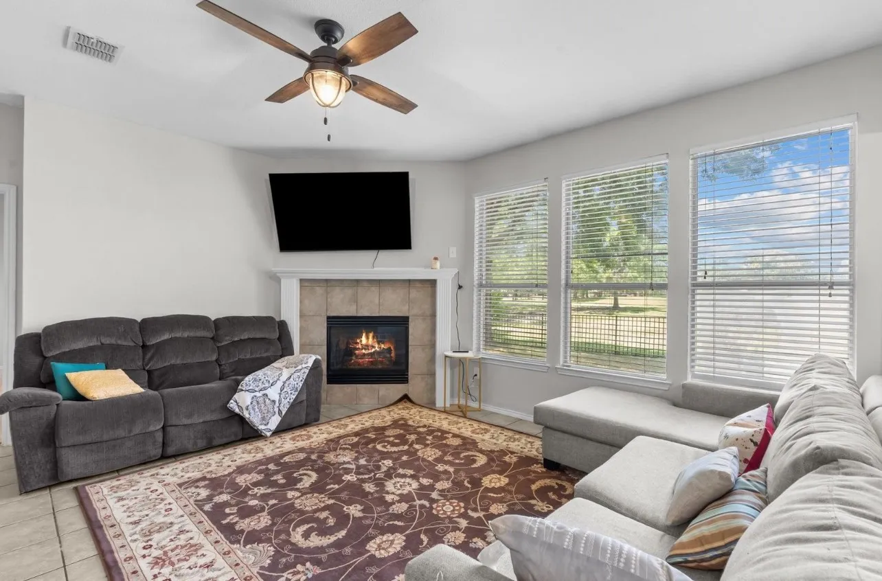Living area with a ceiling fan, tiled fireplace with a white mantel, and three windows offering exterior views