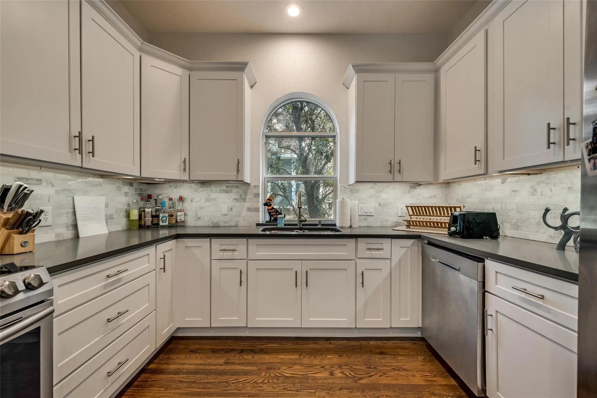 U-shaped kitchen featuring white cabinetry, dark countertops, a tiled backsplash, wood-finish flooring, and an arched window