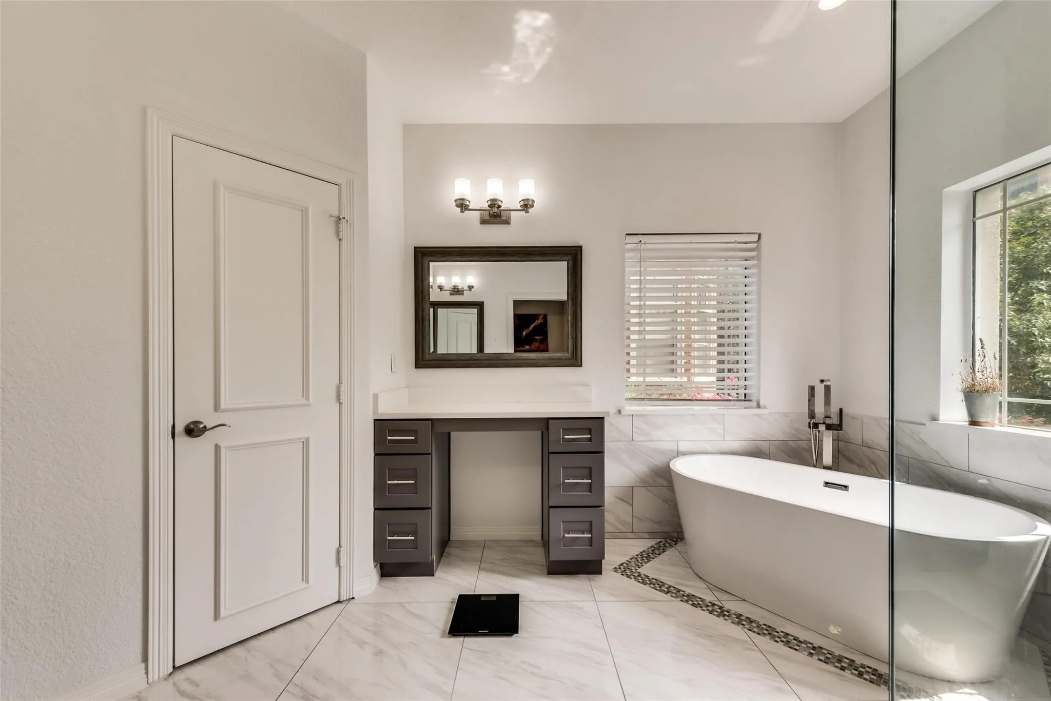 Freestanding bathtub with floor-mounted fixture, glass-enclosed shower, dual-pane window with blinds, and a built-in vanity with countertop