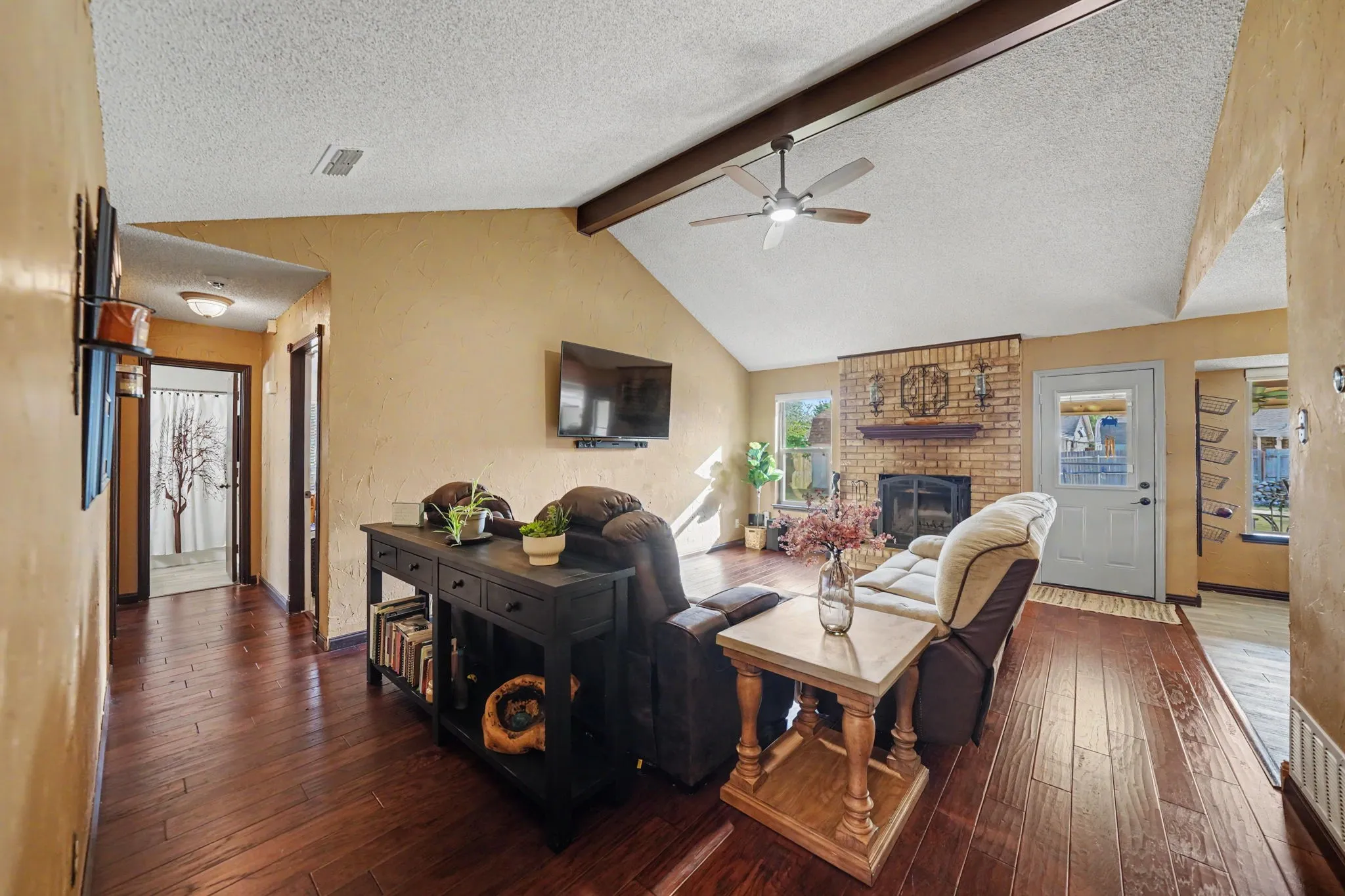 Vaulted ceiling with exposed beams, wood-finish flooring, brick fireplace, ceiling fan, and transom window above door