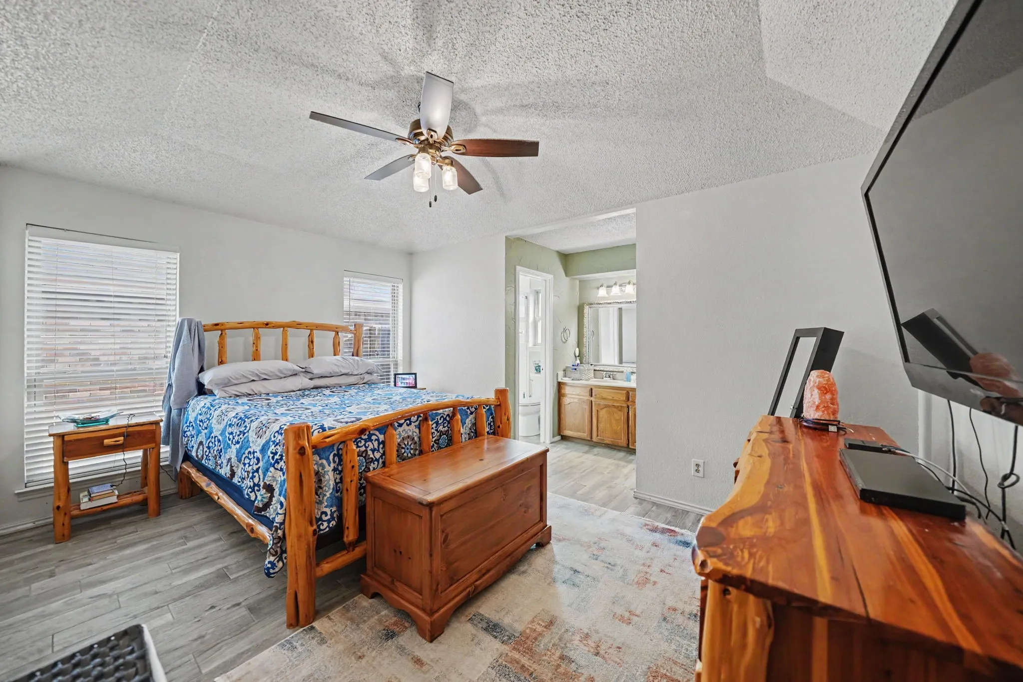 Ceiling fan with integrated lighting, wood-finish flooring, two windows with blinds, recessed lighting in bathroom, and a vanity with dual sinks