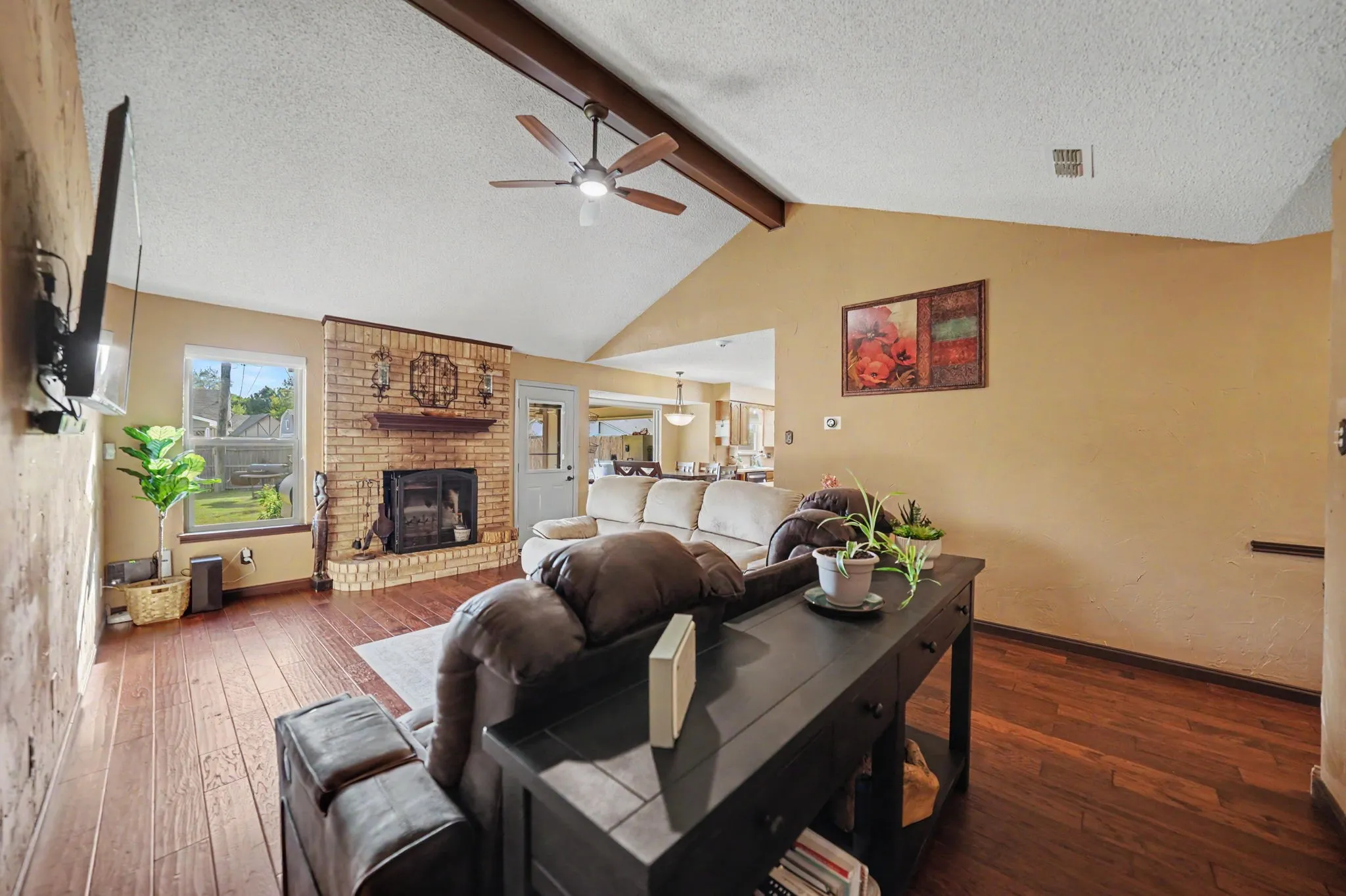 Vaulted ceiling with exposed wood beam, brick fireplace with mantel, wood-finish flooring, and a ceiling fan with integrated lighting