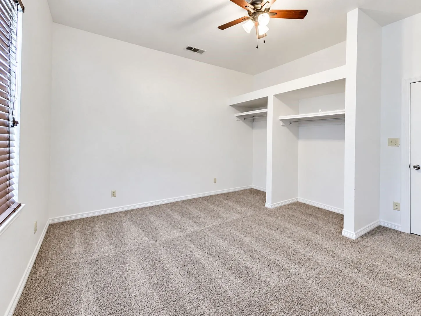 Neutral tone wall paint, textured carpeting, built-in closet shelving, window with horizontal blinds, and a ceiling fan