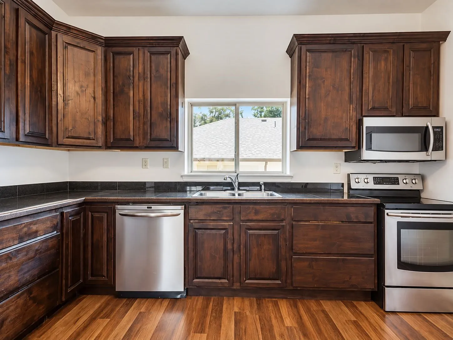 Dark wood cabinetry with crown molding, stainless steel appliances, black countertops, wood-finish flooring, and a double basin sink