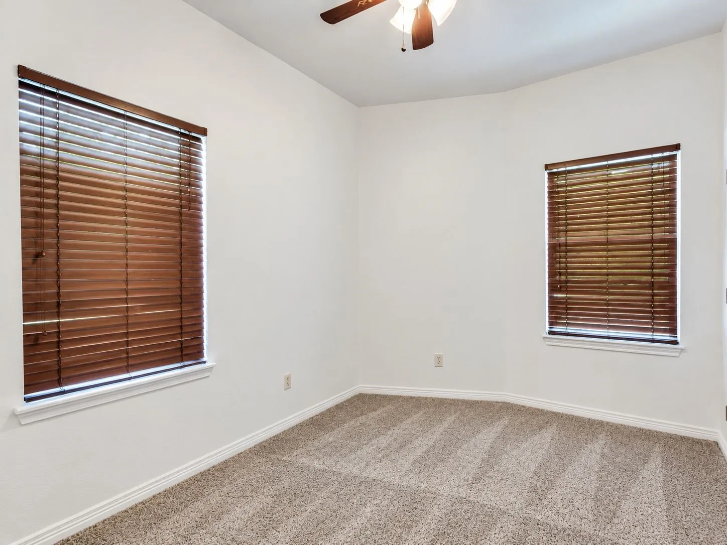 Neutral-toned carpet flooring, white walls, two windows with wood-finish blinds, and a ceiling fan with integrated lighting