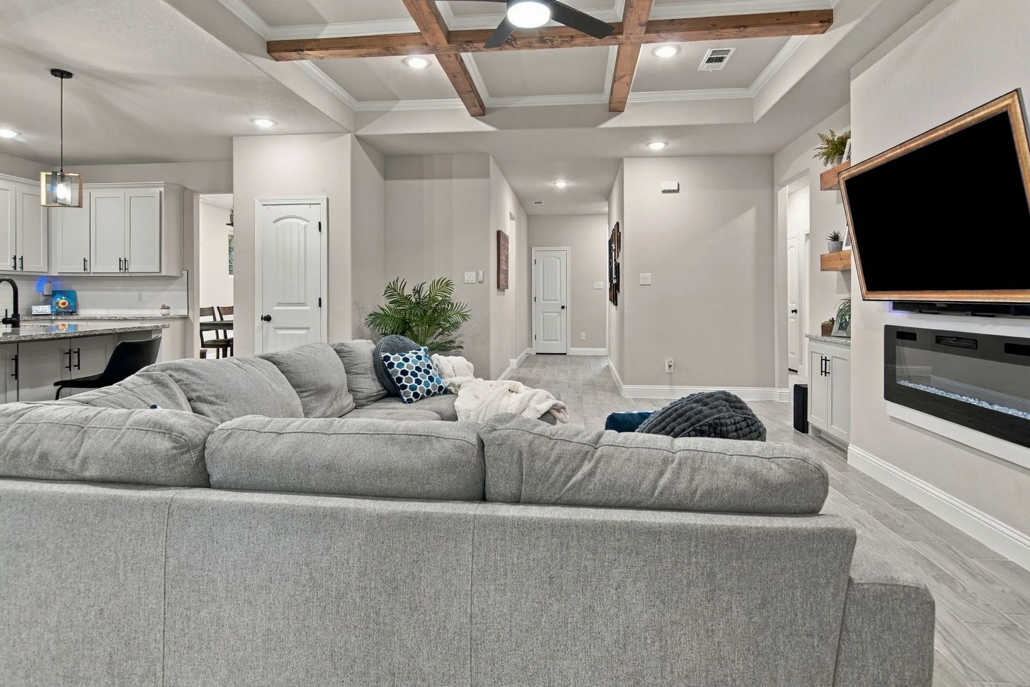 Tray ceiling with exposed wood beams, recessed lighting, wood-finish flooring, built-in electric fireplace, and kitchen with white cabinetry