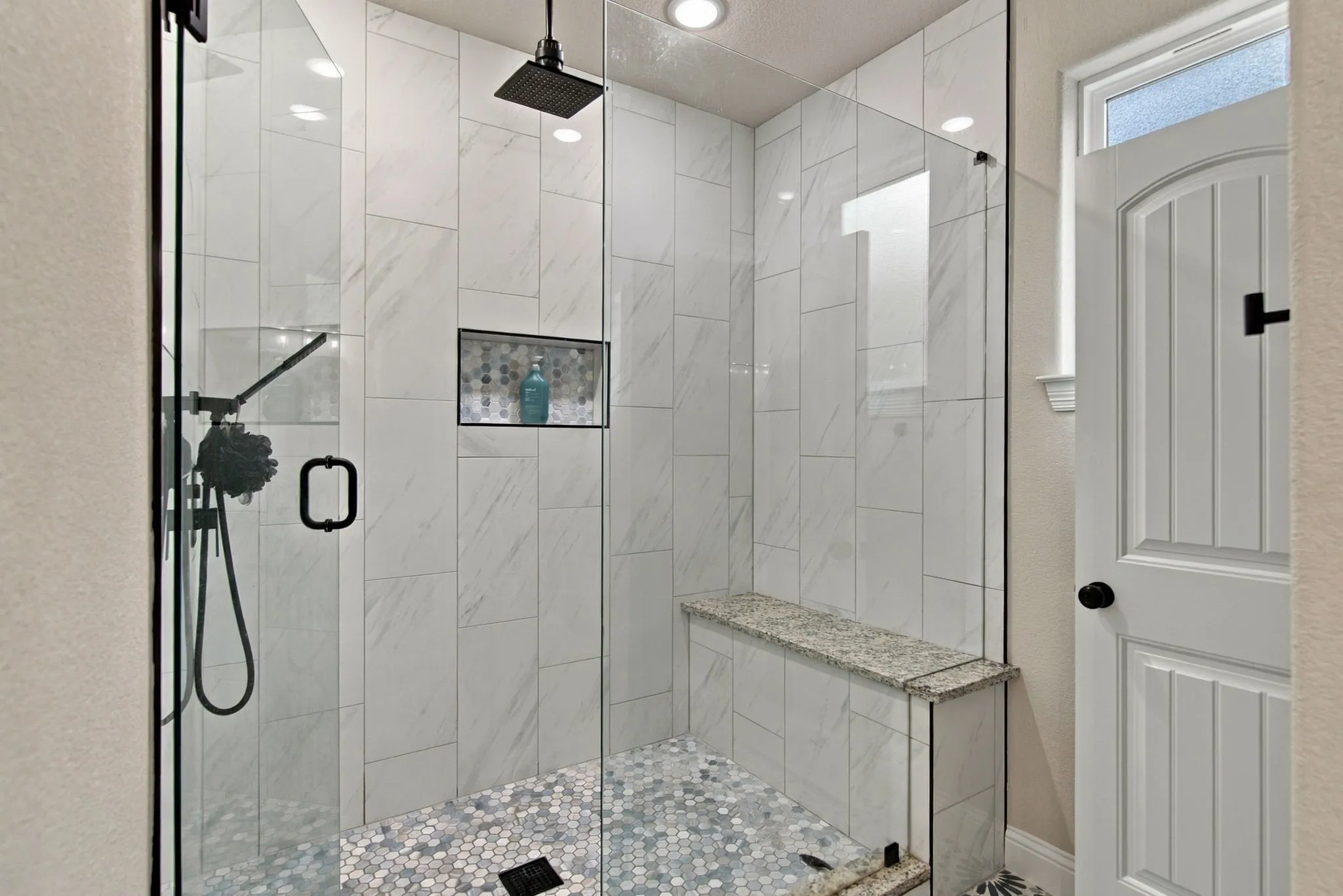 Frameless glass shower enclosure with matte black fixtures, floor-to-ceiling tile, built-in shower bench, and recessed niche