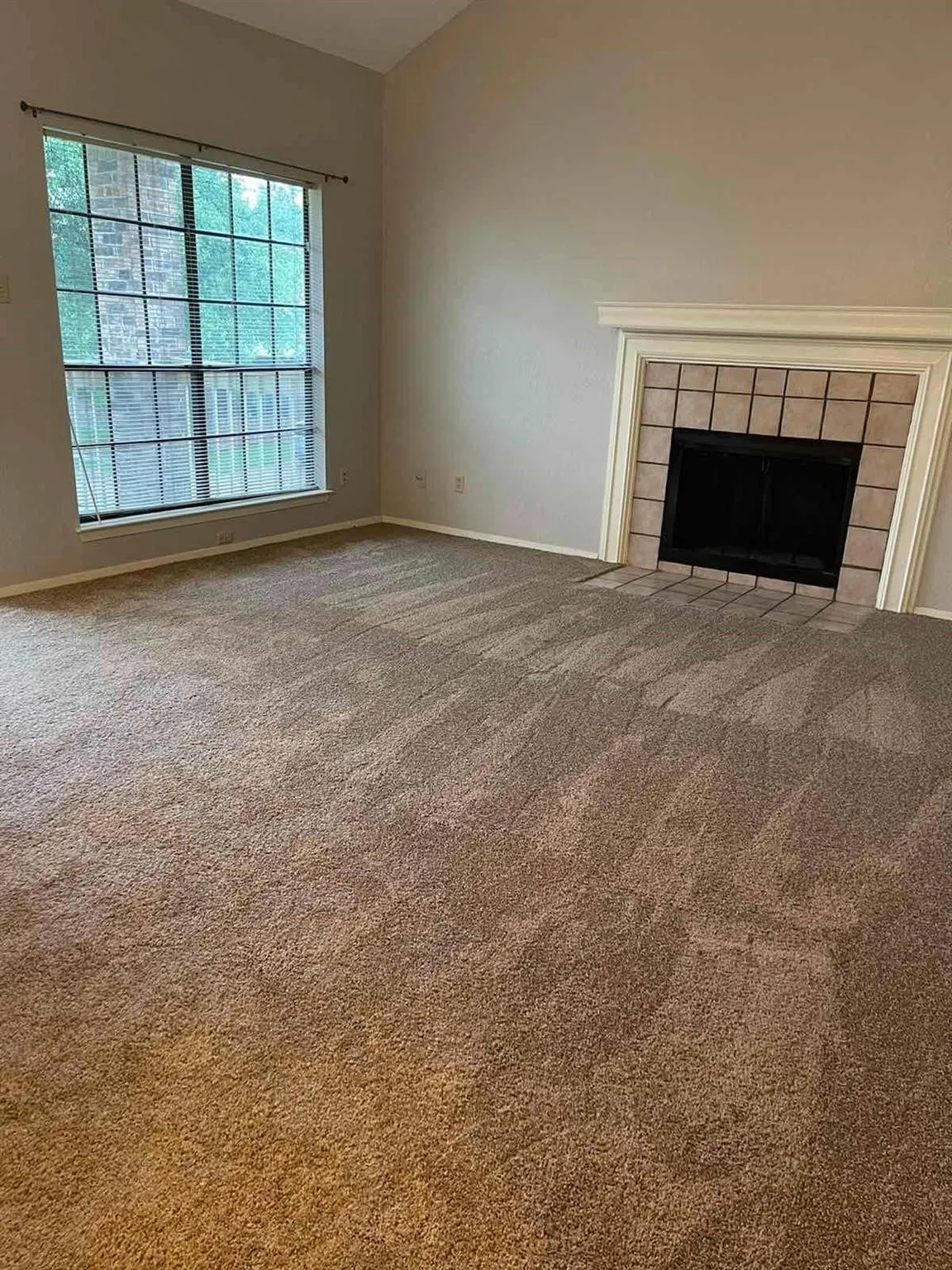 Vaulted ceiling living area with a large multi-pane window, carpet flooring, and a tiled fireplace with a mantel