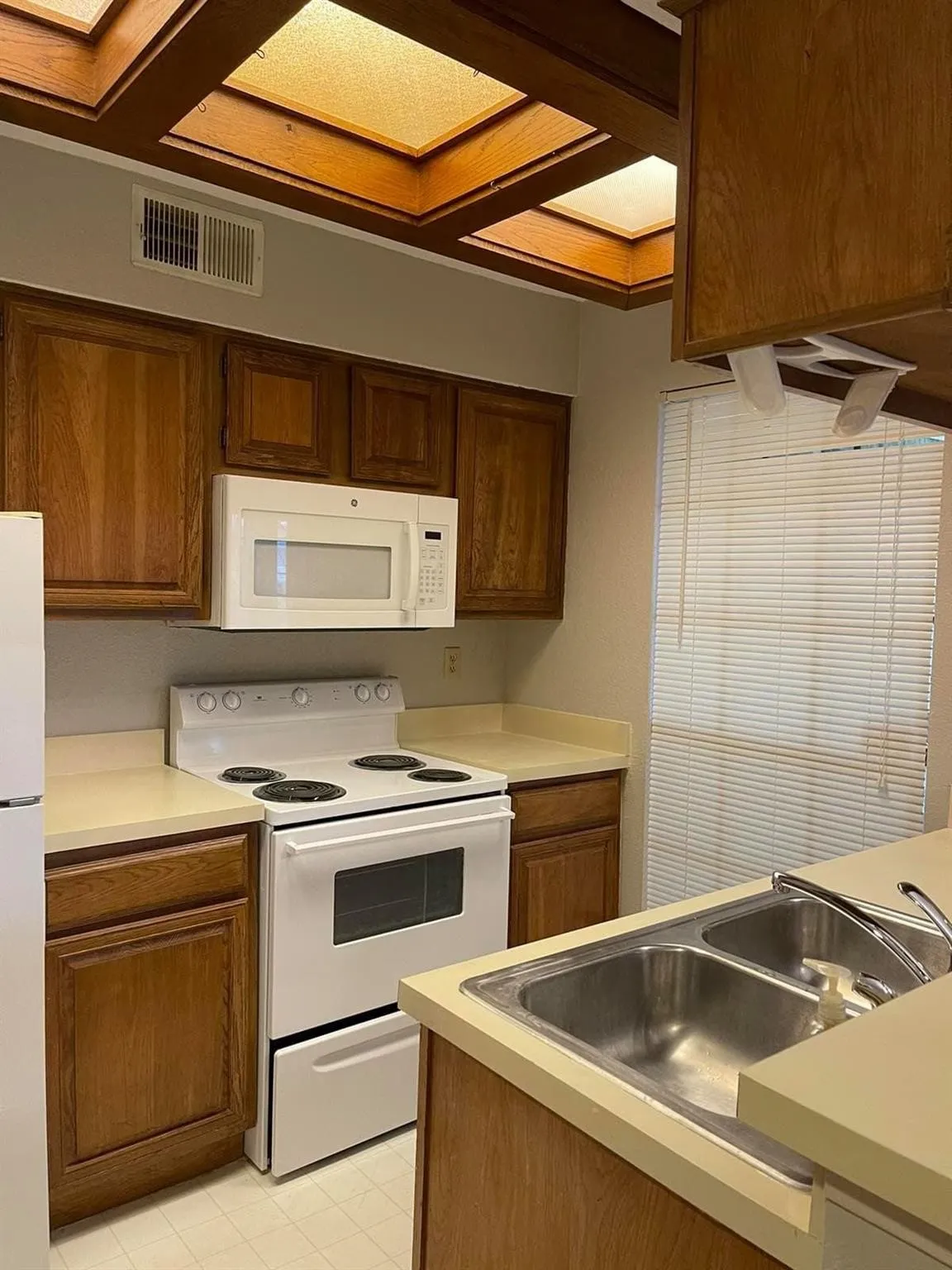 Kitchen featuring wood cabinetry, white appliances, dual basin stainless steel sink, light-toned countertops, and ceiling skylights