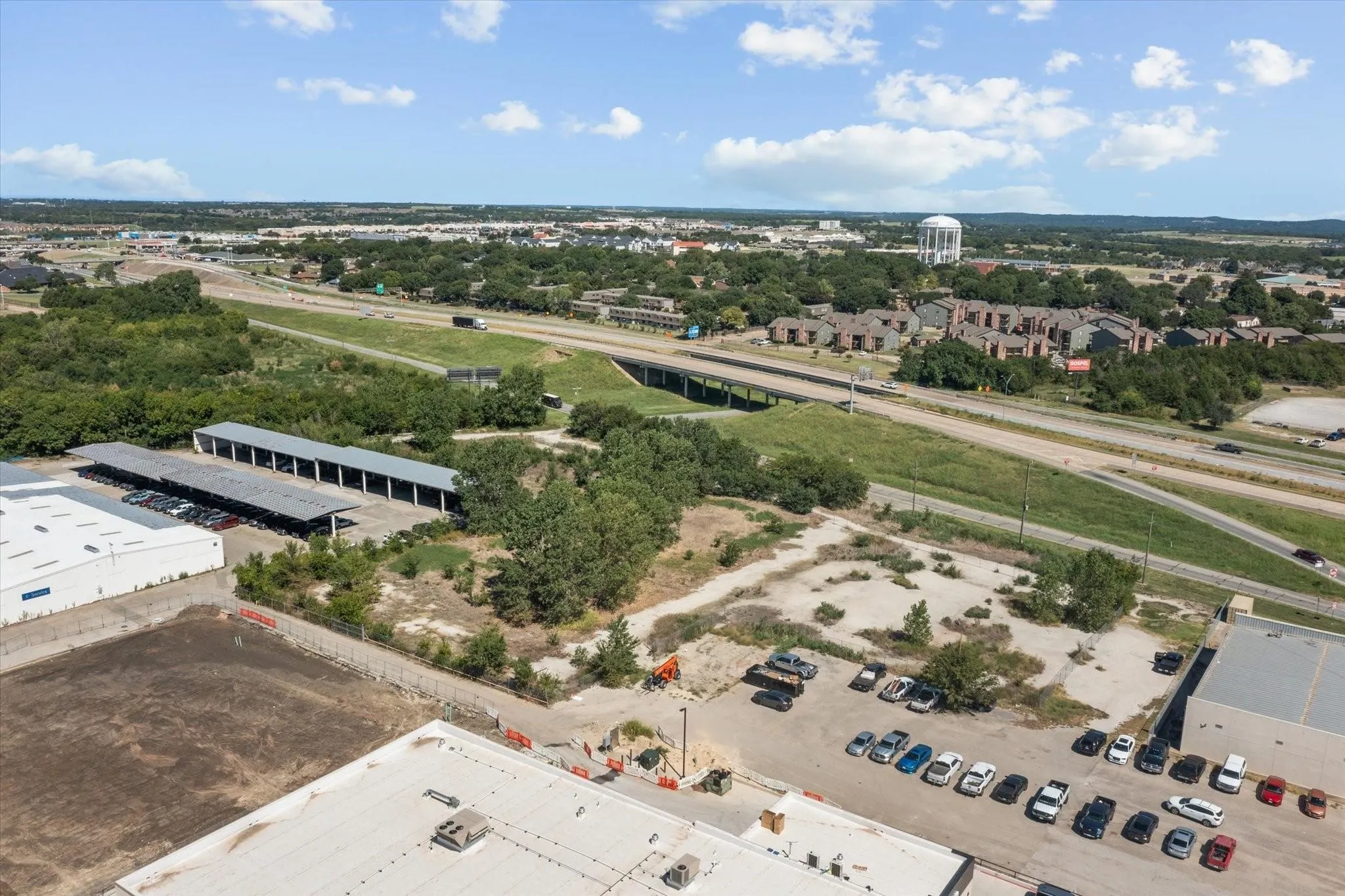 Expansive land parcel featuring a paved parking area, adjacent to a major roadway, with surrounding commercial structures and mature trees