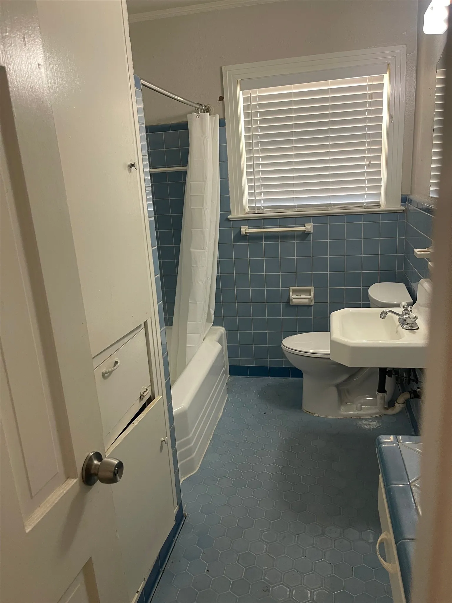 Built-in linen cabinetry, window with blinds, subway-tiled bathtub/shower combination, pedestal sink, and blue hexagonal floor tiling
