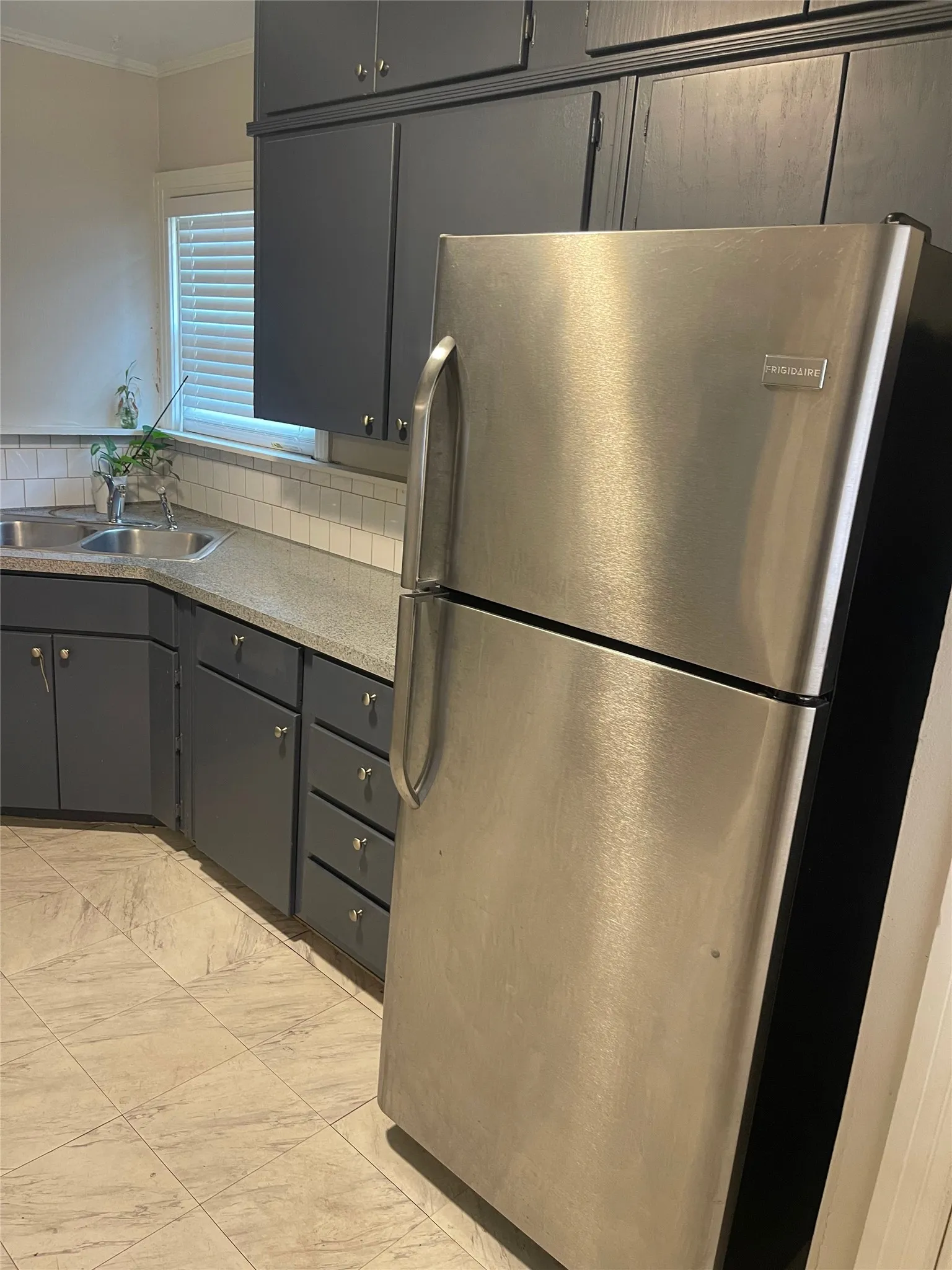Kitchen featuring dark cabinetry, light-toned countertops, a stainless steel refrigerator, a double basin sink, and a tile backsplash