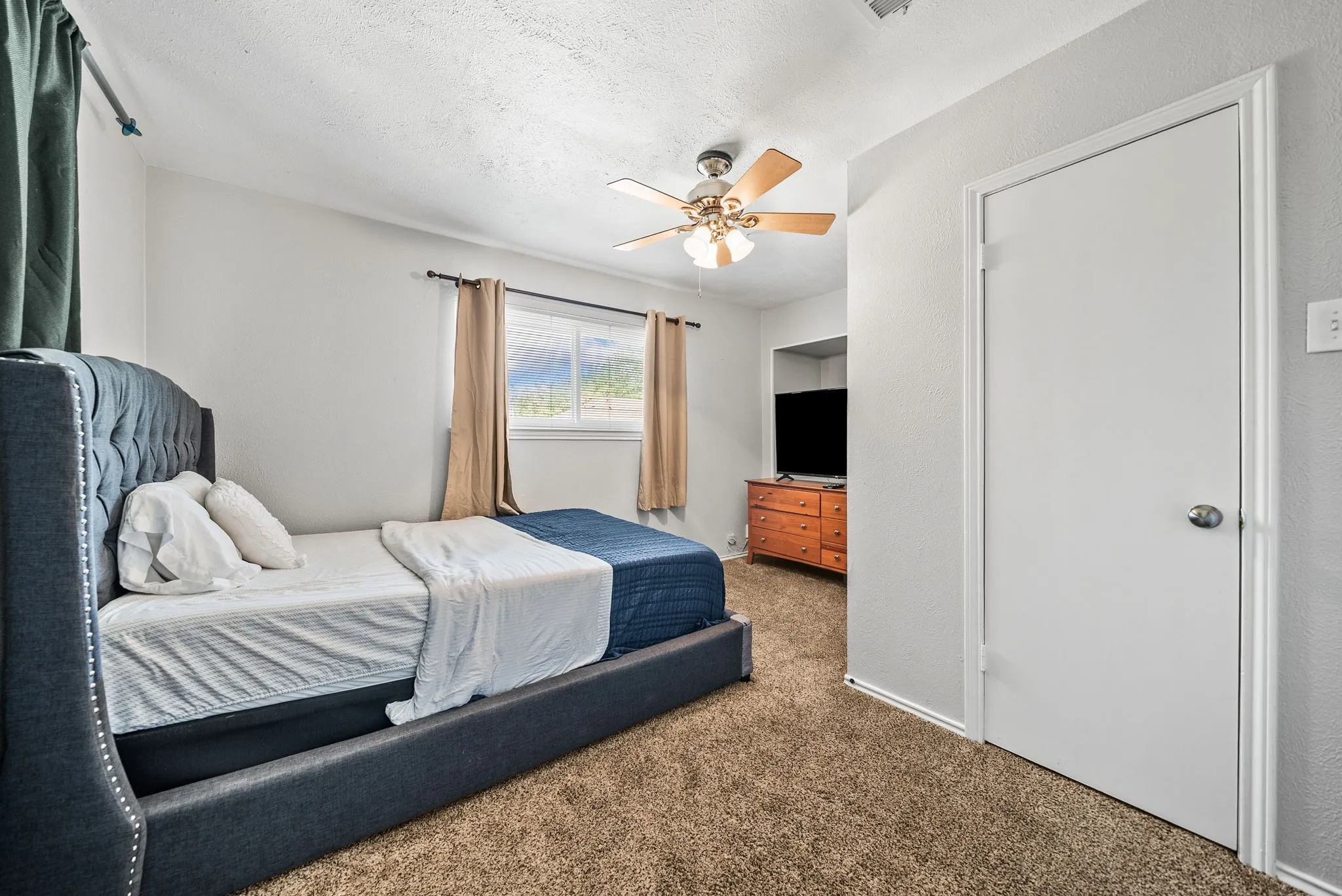 Neutral wall paint, textured ceiling, window with exterior scenery, ceiling fan with light fixture, and carpet flooring
