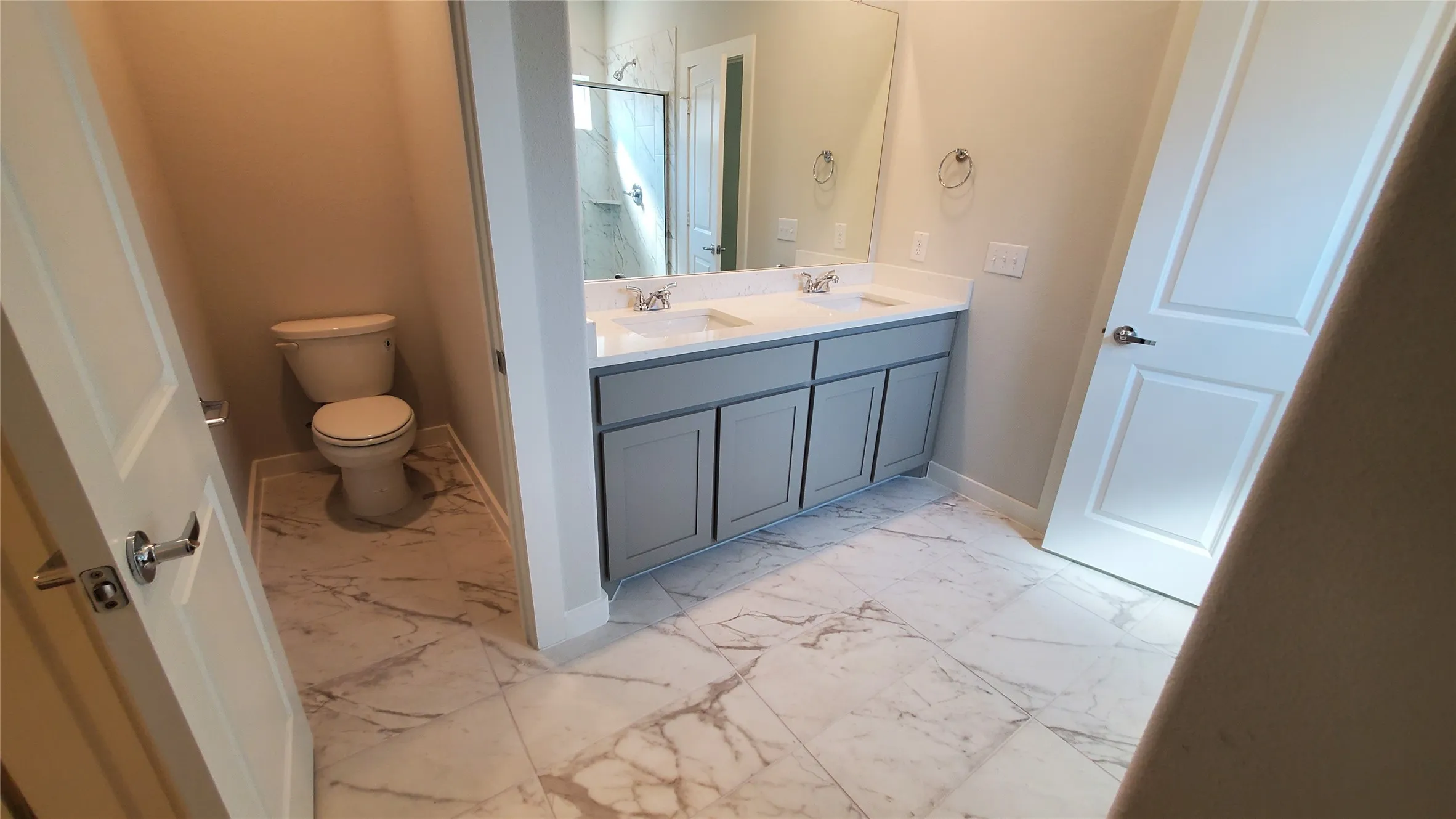 Dual vanity with undermount sinks and light gray cabinetry, white marble-look tile flooring, and a glass-enclosed shower
