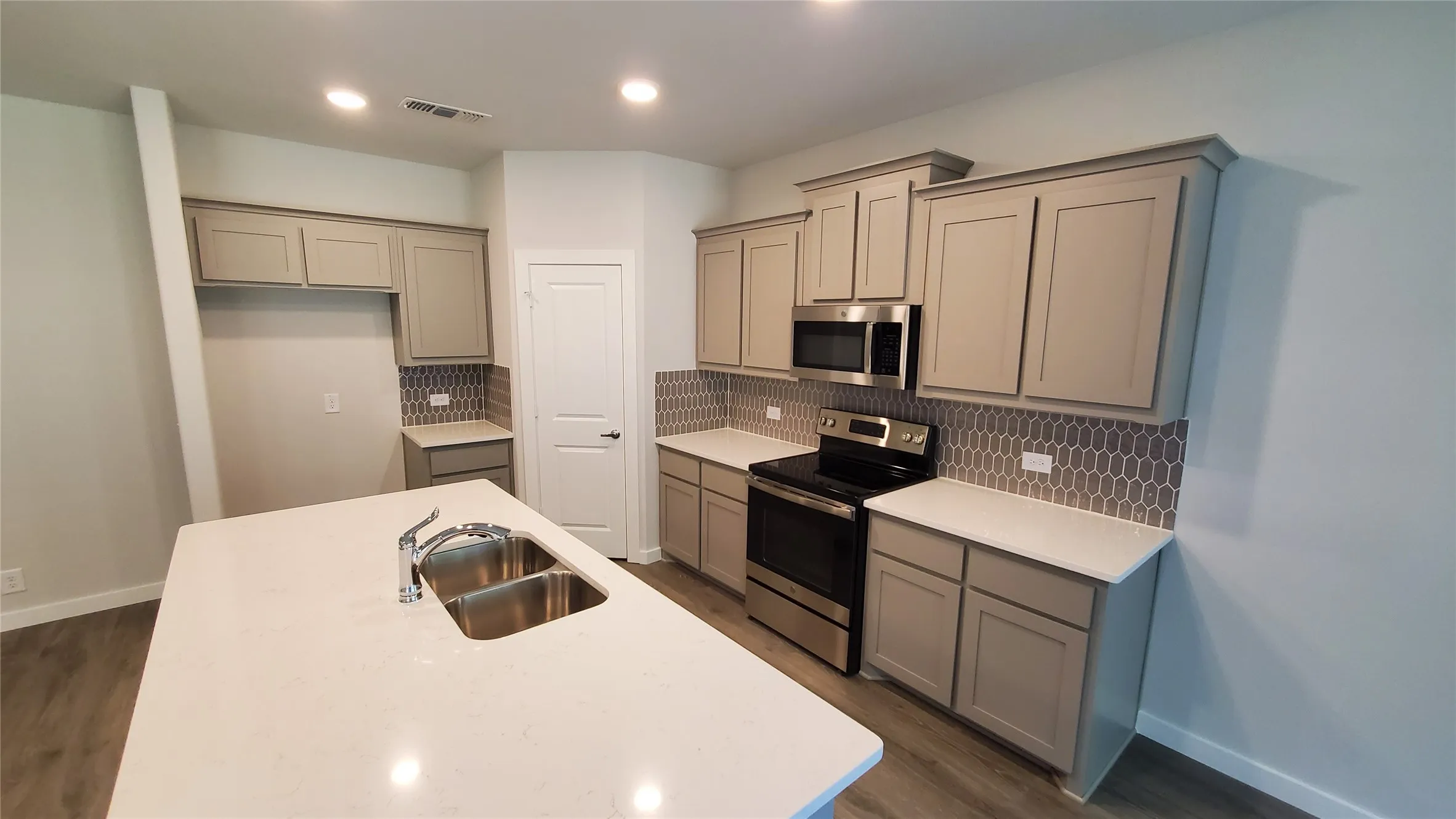 Kitchen featuring an island with a double basin sink, light-toned cabinetry, light countertops, wood-finish flooring, and a patterned tile backsplash