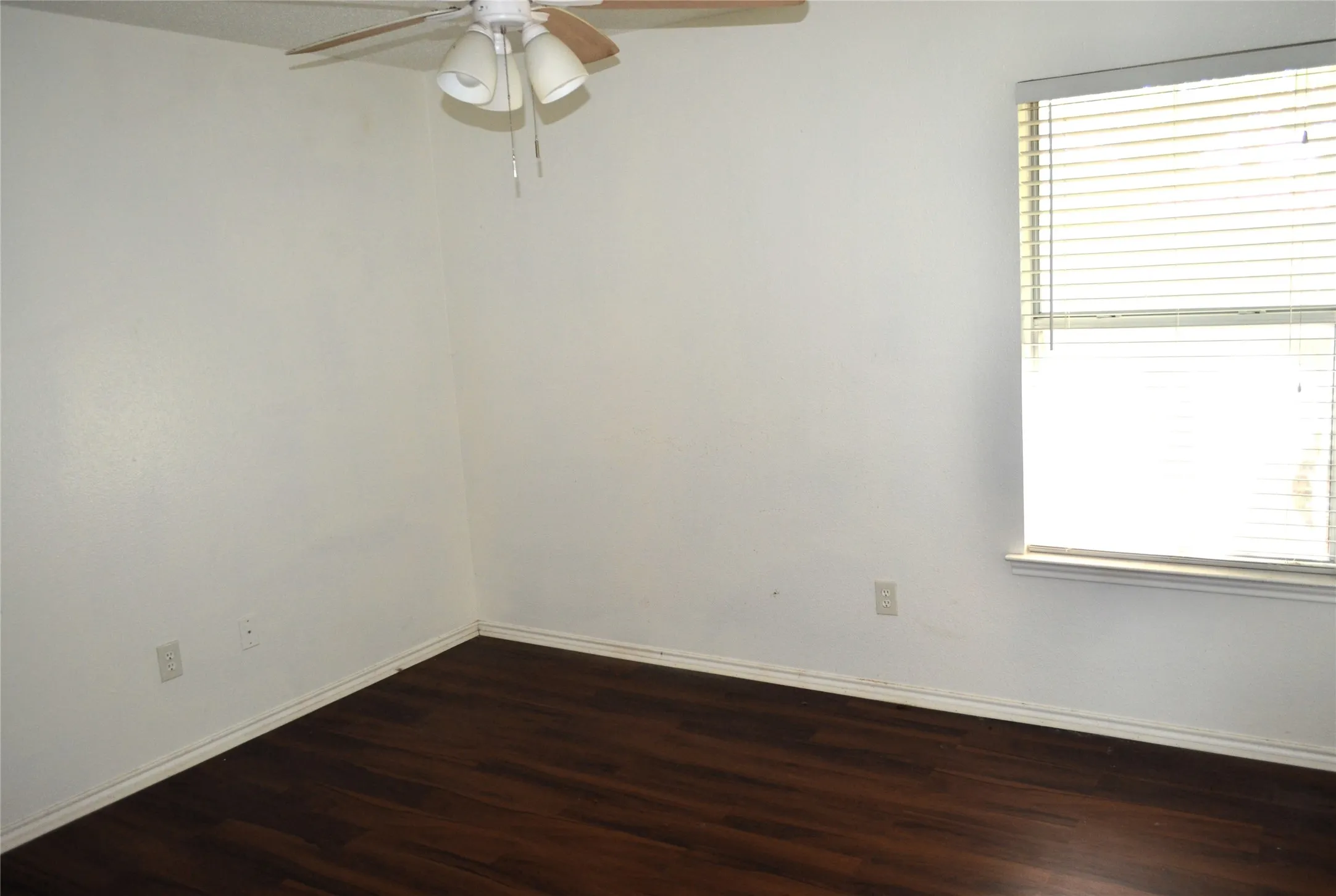 Wood-finish flooring with white baseboards, window with blinds, and a ceiling fan with integrated lighting
