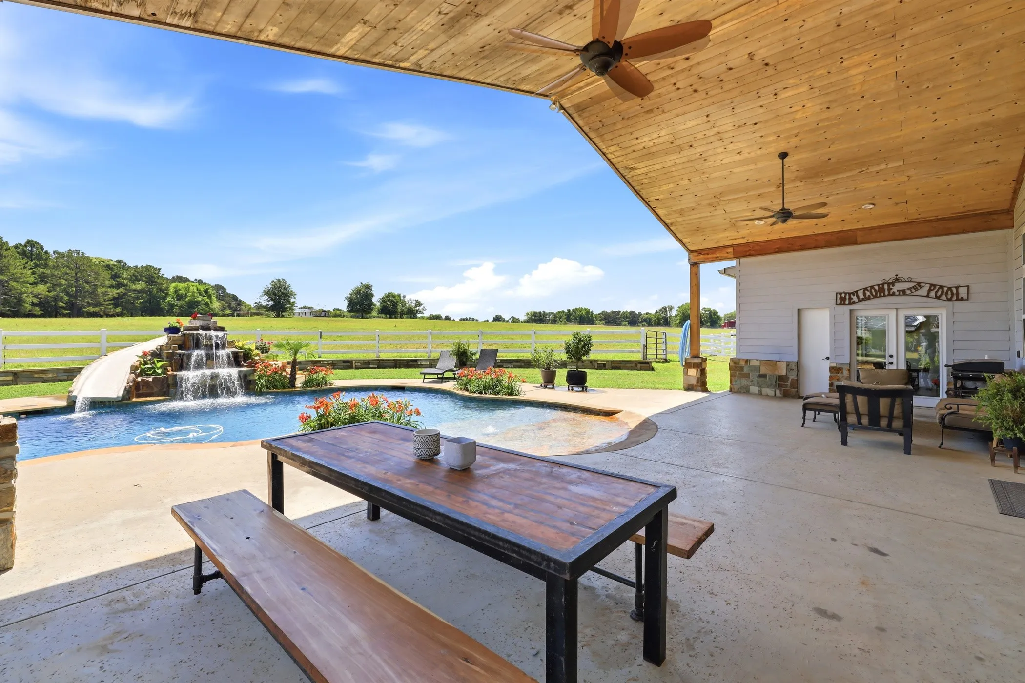 Expansive concrete patio with a wooden ceiling, integrated pool with a rock waterfall feature, and a built-in pool slide