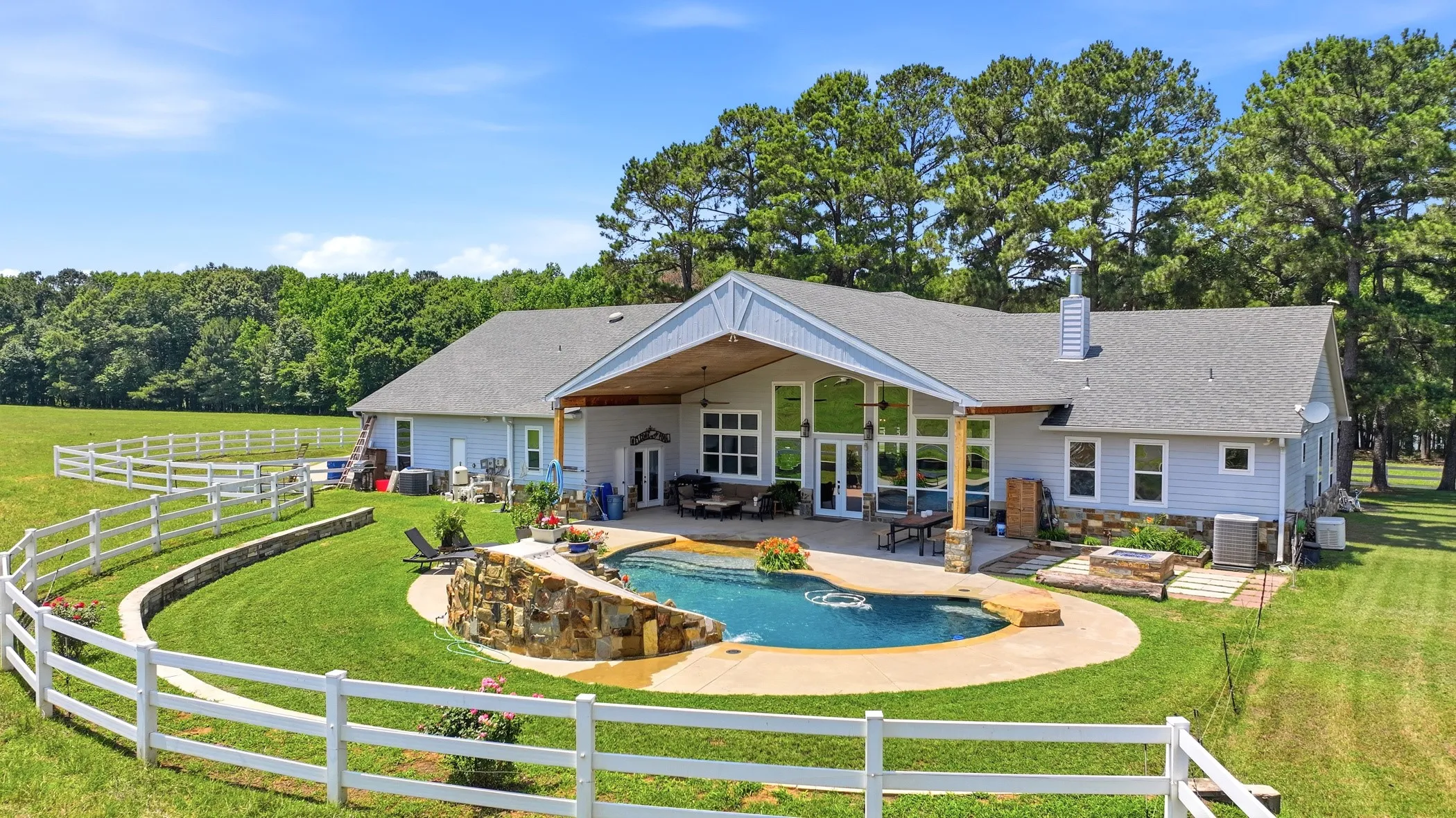 Spacious property featuring a large inground pool with rock detailing, extensive patio area, covered outdoor living space, and a white picket fence
