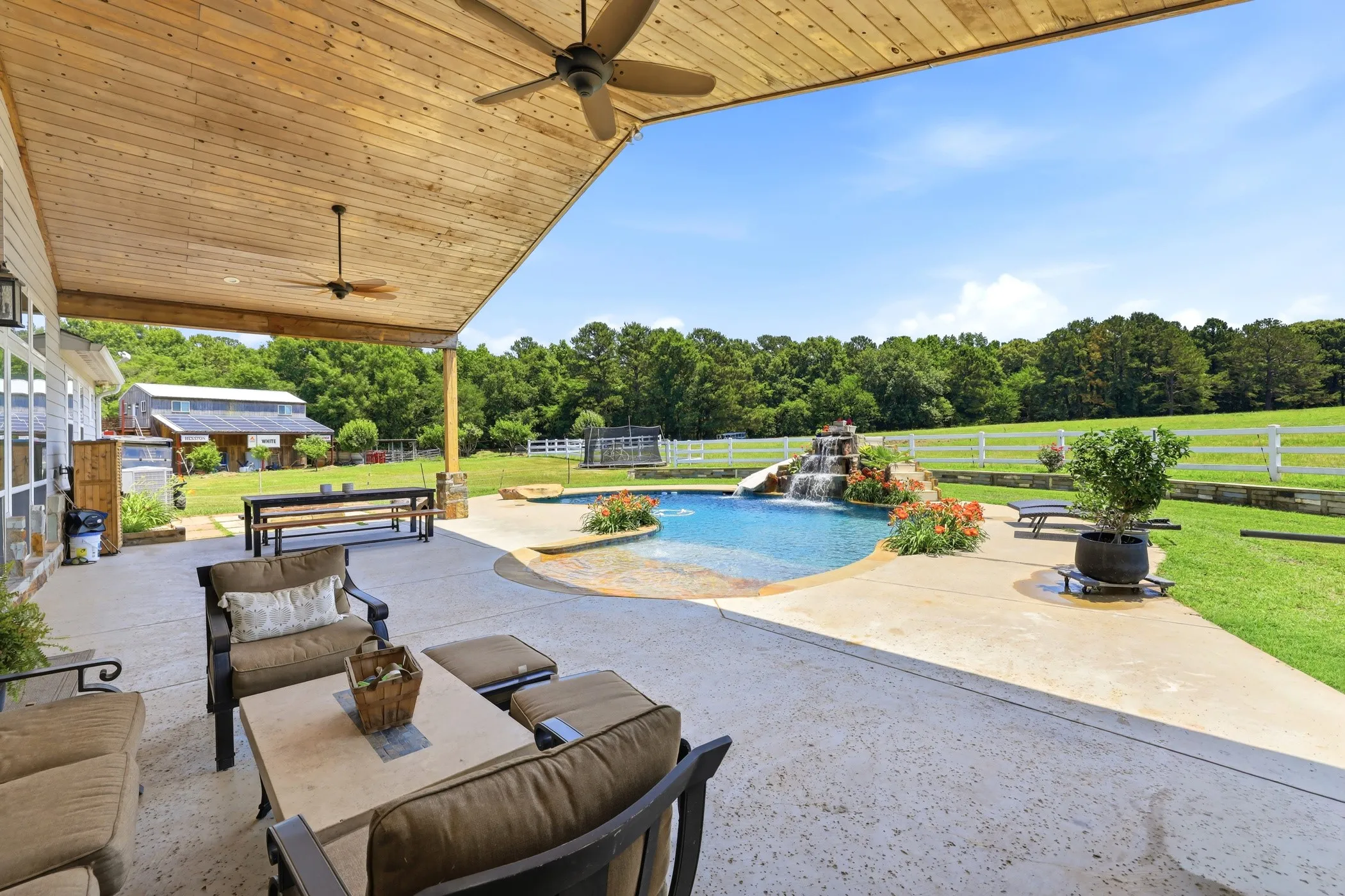 Covered patio with wood-finish ceiling and ceiling fans, leading to a custom-shaped pool with a rock waterfall feature, concrete decking, and expansive lawn area