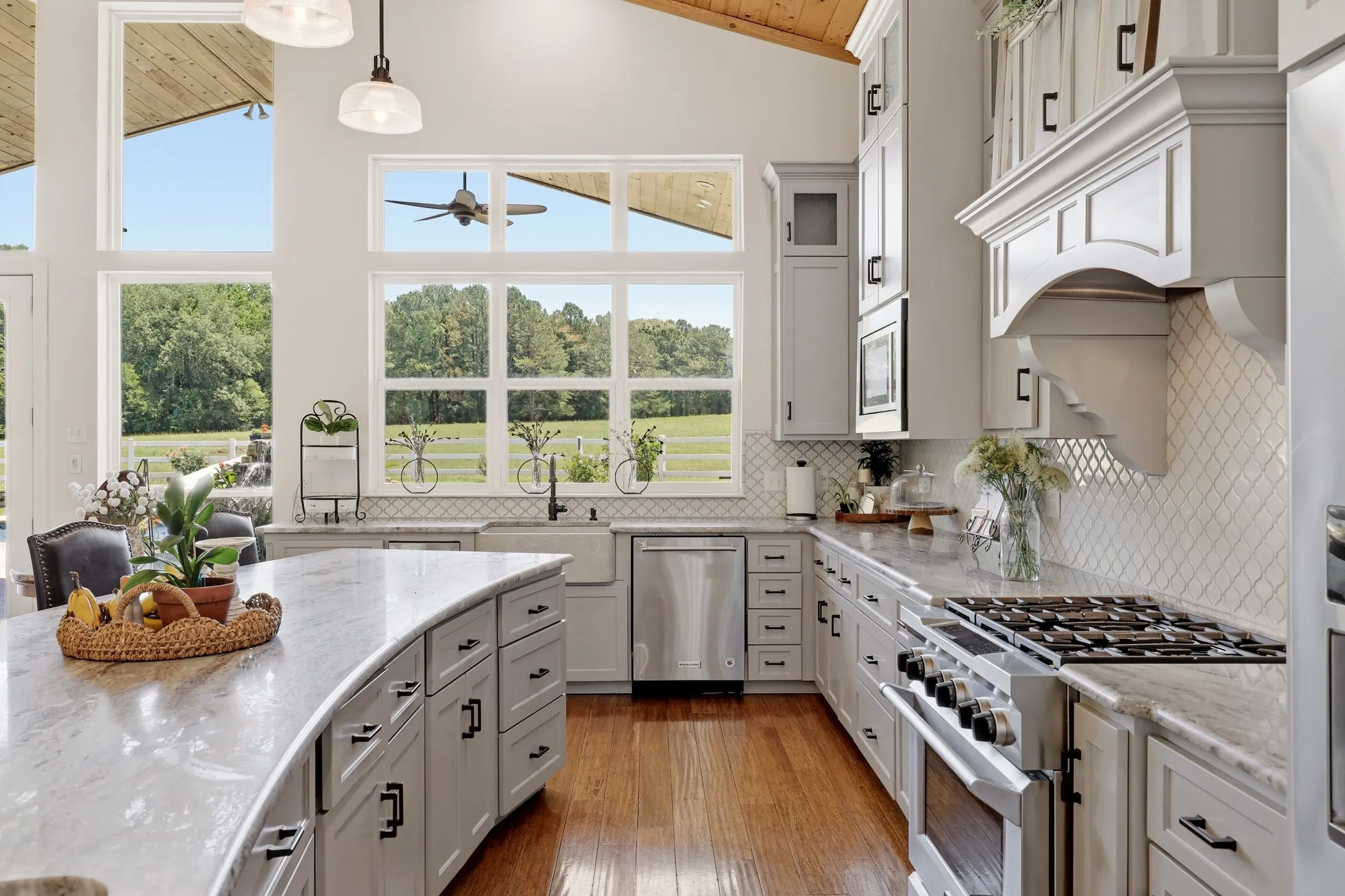 Vaulted wood-finish ceiling, expansive window wall, curved kitchen island with stone countertop, stainless steel appliances, and custom range hood