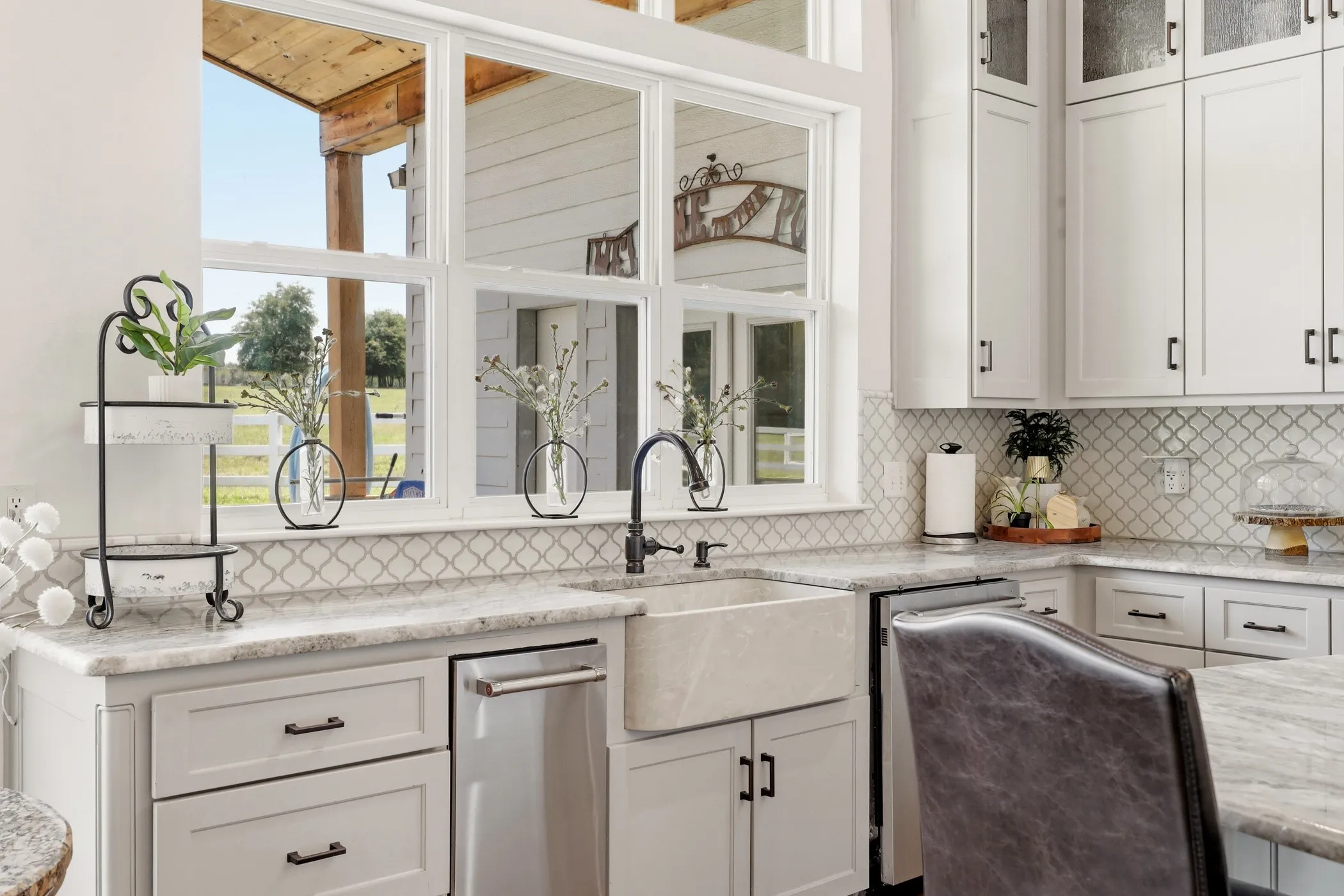 White shaker-style cabinetry with dark hardware, stone countertops, arabesque tile backsplash, farmhouse sink with a gooseneck faucet, and a large window overlooking an outdoor patio