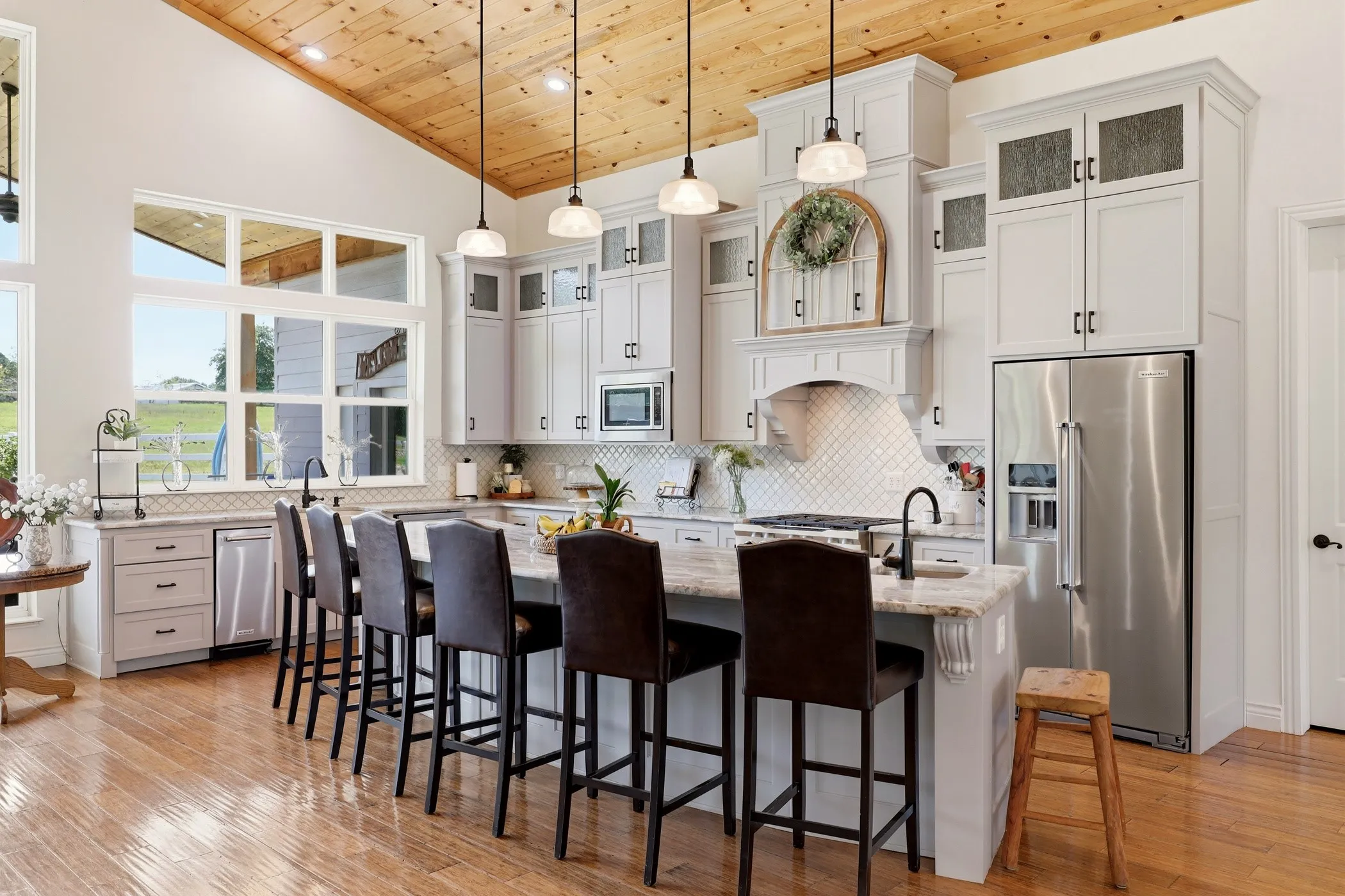 Vaulted wood-paneled ceiling, expansive window array, light-toned cabinetry with glass inserts, large center island with seating, and wood-finish flooring
