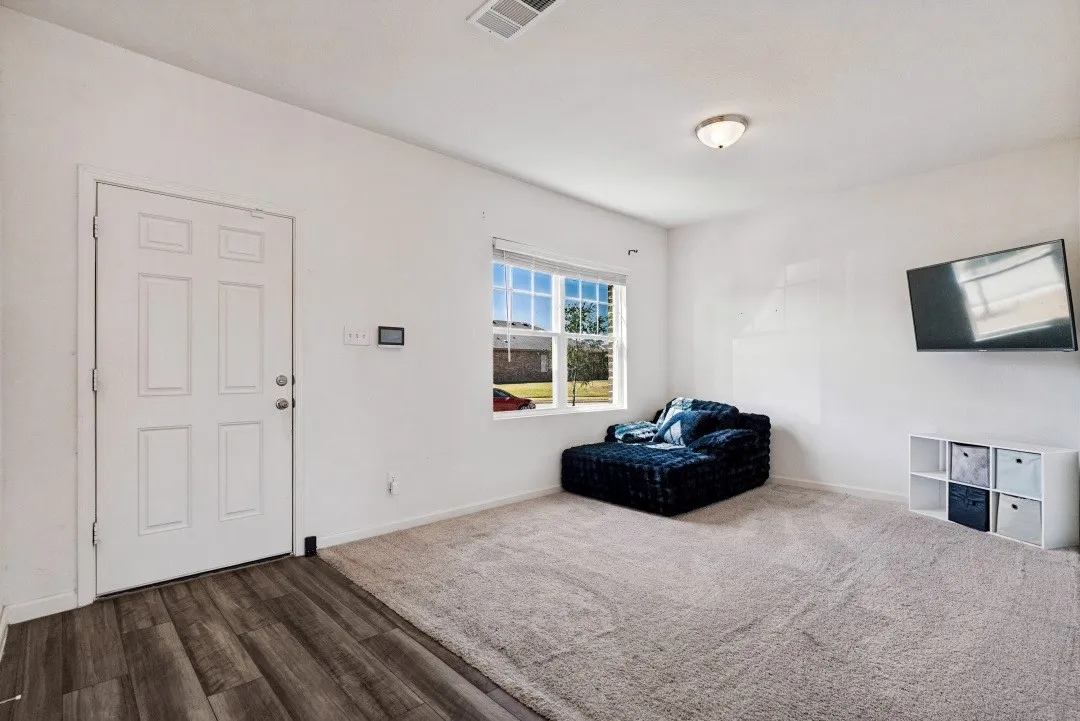 White paneled door, wood-finish flooring, light neutral carpeting, recessed wall-mounted screen, and a flush-mount ceiling light