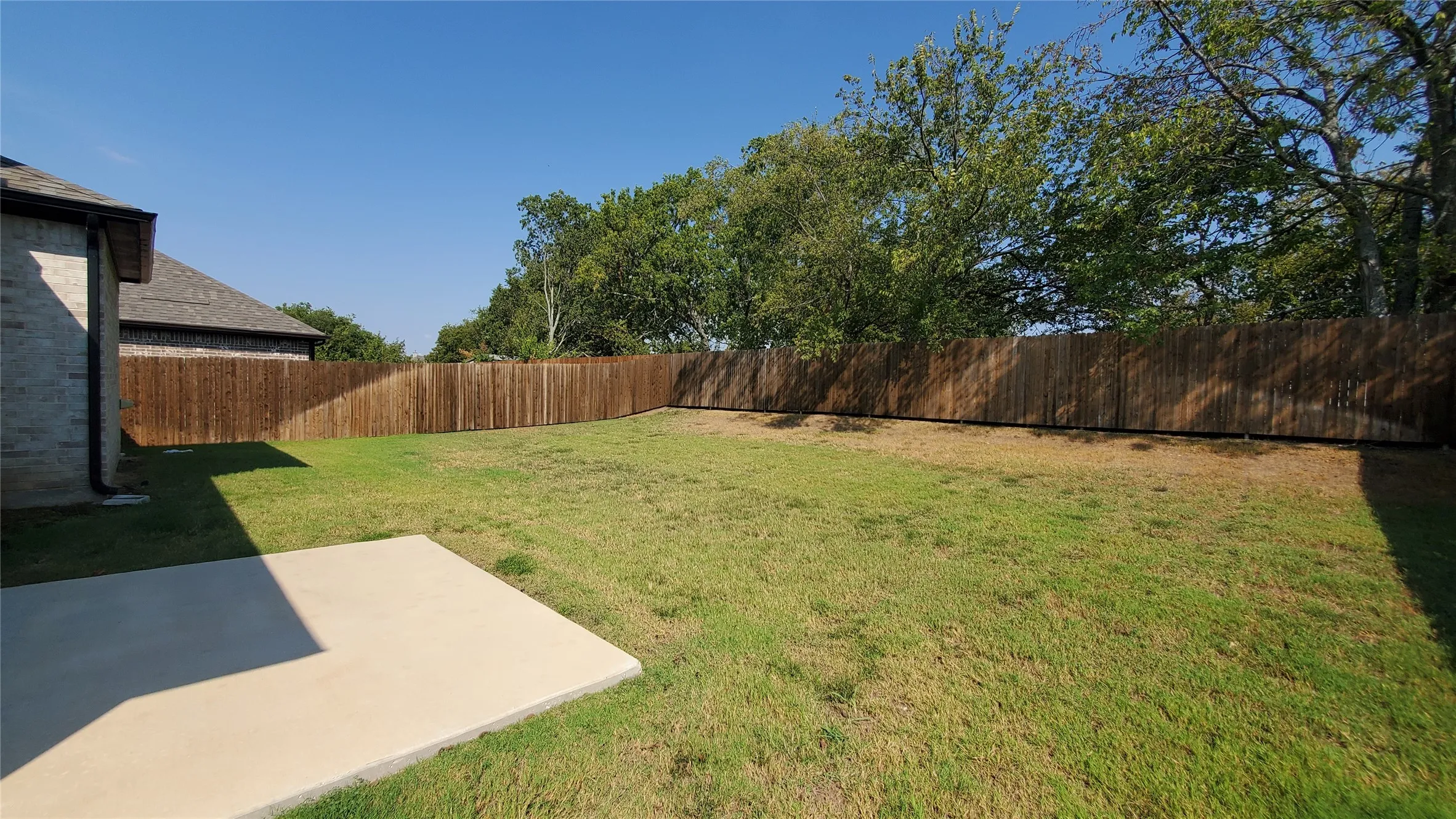 Expansive grassy yard with a wooden privacy fence, featuring a concrete patio slab and a stone-clad exterior