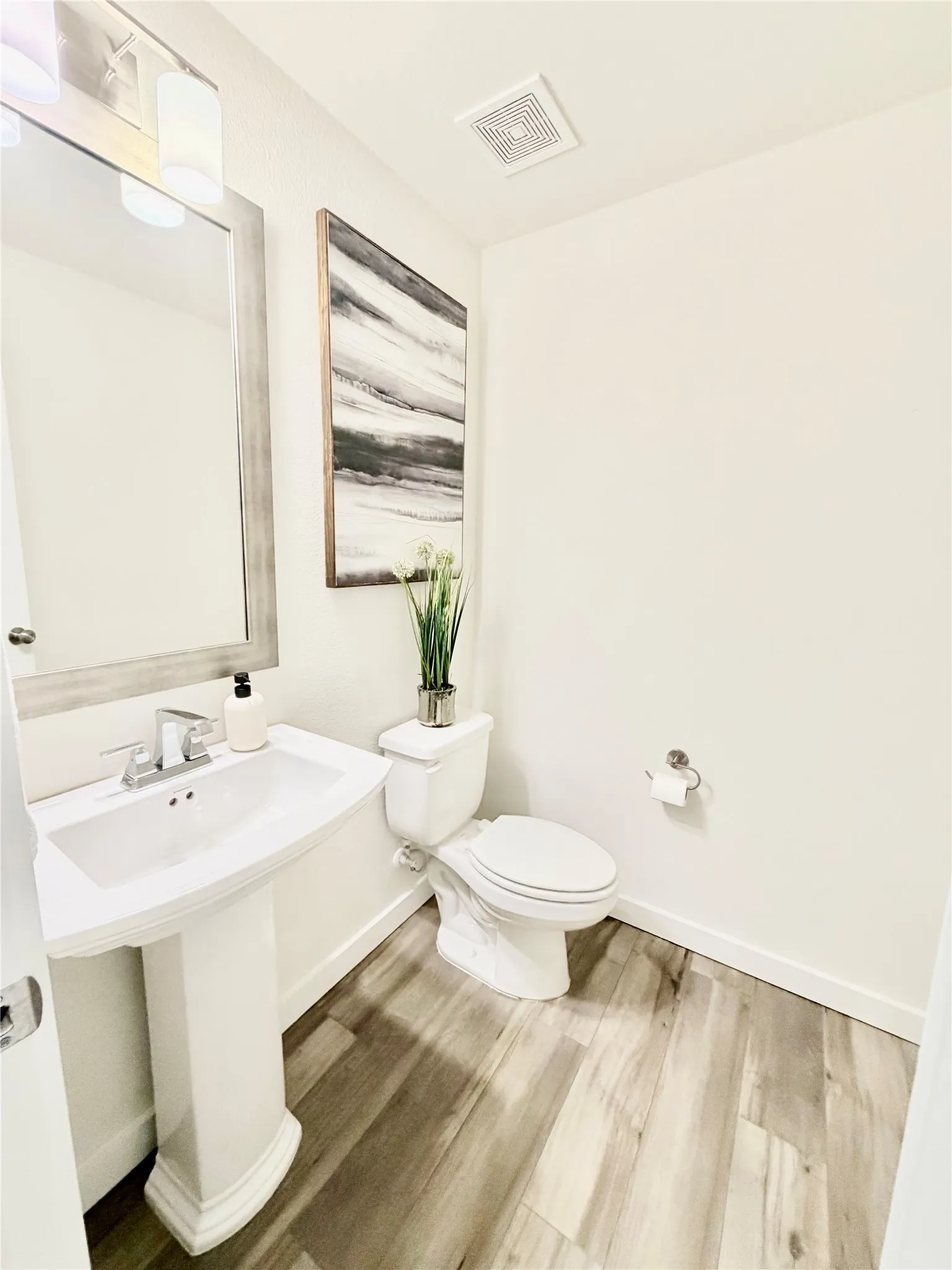 Pedestal sink with chrome faucet, a white toilet, wood-finish flooring, and a rectangular mirror with an integrated light fixture