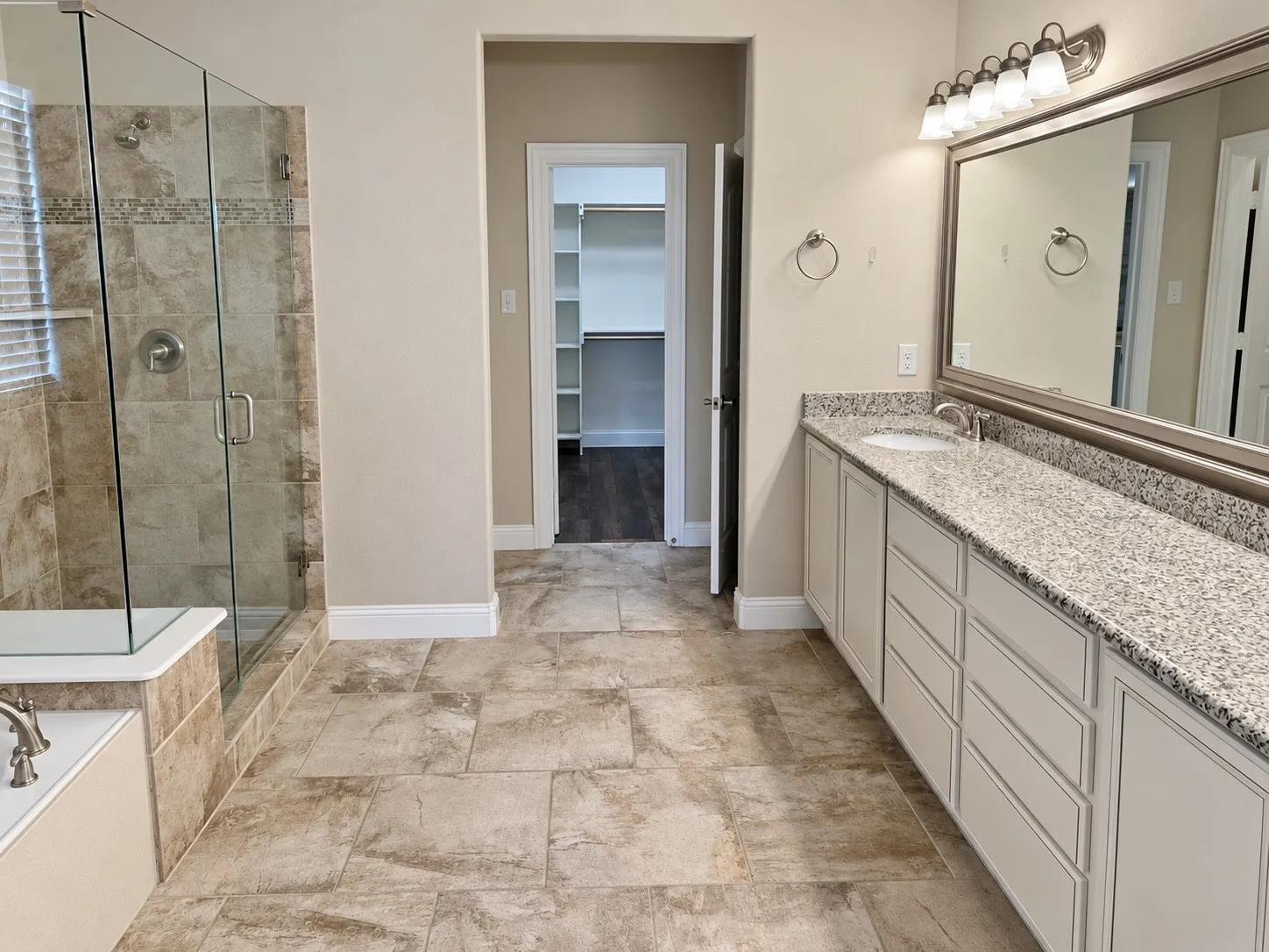 Bathroom featuring a dual-sink vanity with granite countertops, a framed mirror, a glass-enclosed shower with tile surround, and large format floor tiling