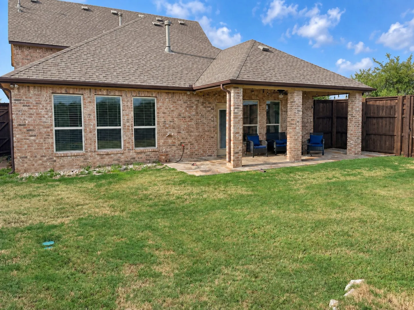 Brick exterior with a covered patio, featuring a brown shingle roof, multiple windows, and a wood privacy fence