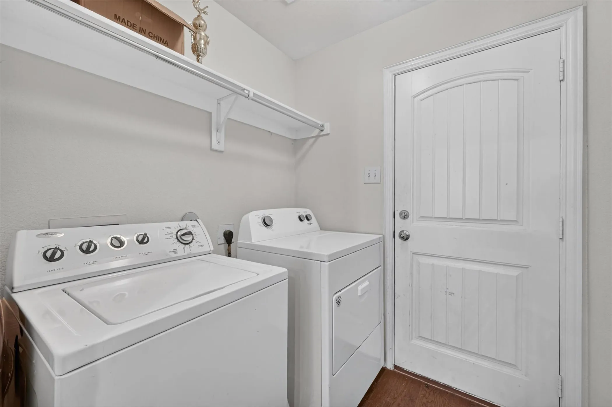 Utility room featuring white walls, durable wood-finish flooring, a white paneled door, and a white storage shelf